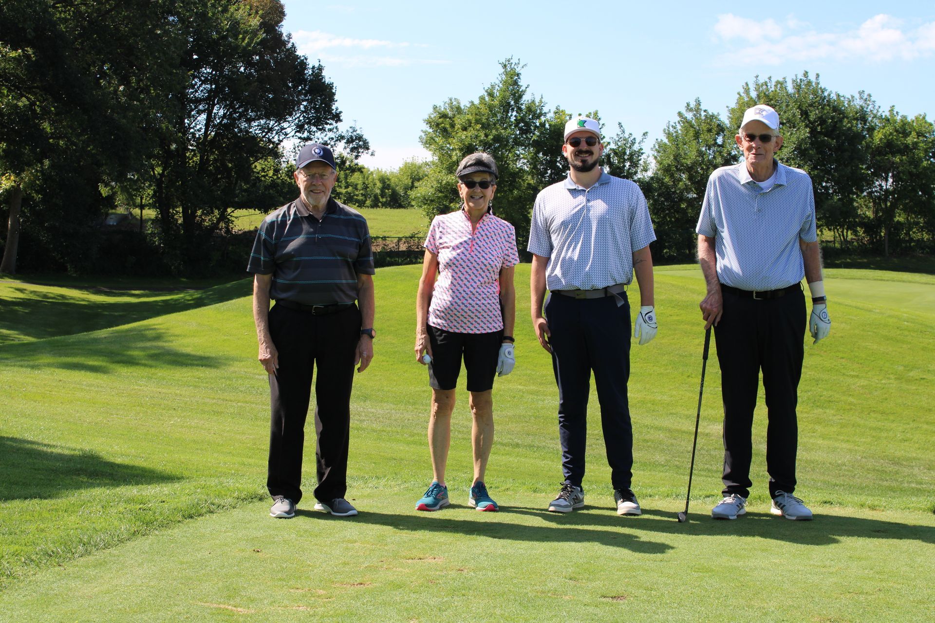 Four people stand in a line on a grassy golf course wearing golf attire, holding clubs, and smiling on a sunny day.