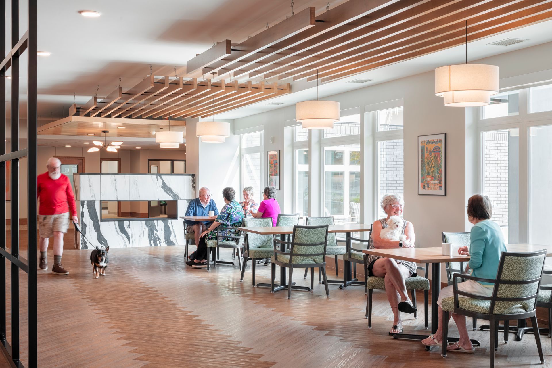 A bright, modern common area with people dining at tables, a person walking a dog, and light wood-slat ceiling accents.