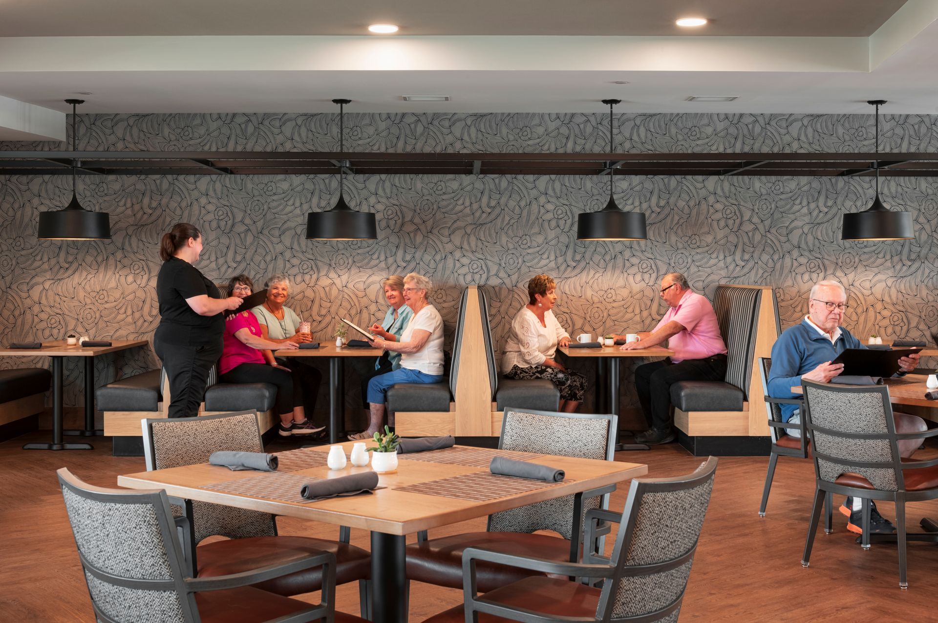 A server takes orders from diners in booths and at tables inside a modern restaurant with pendant lighting.