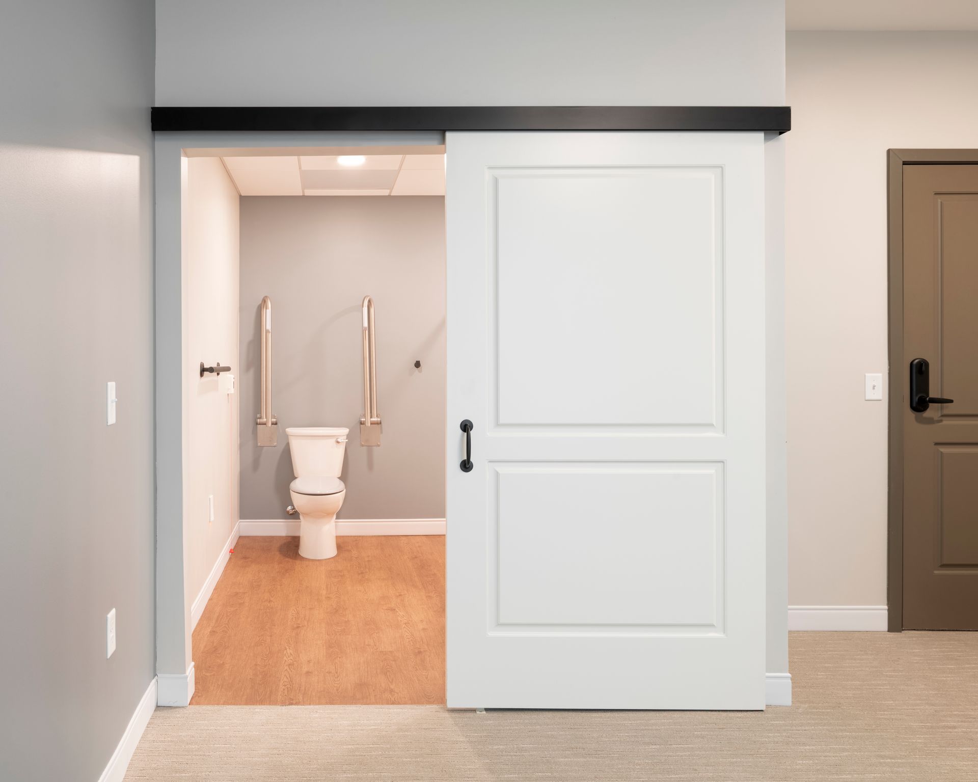 A white sliding barn door opens to a bathroom featuring a toilet with wall-mounted grab bars and light wood flooring.
