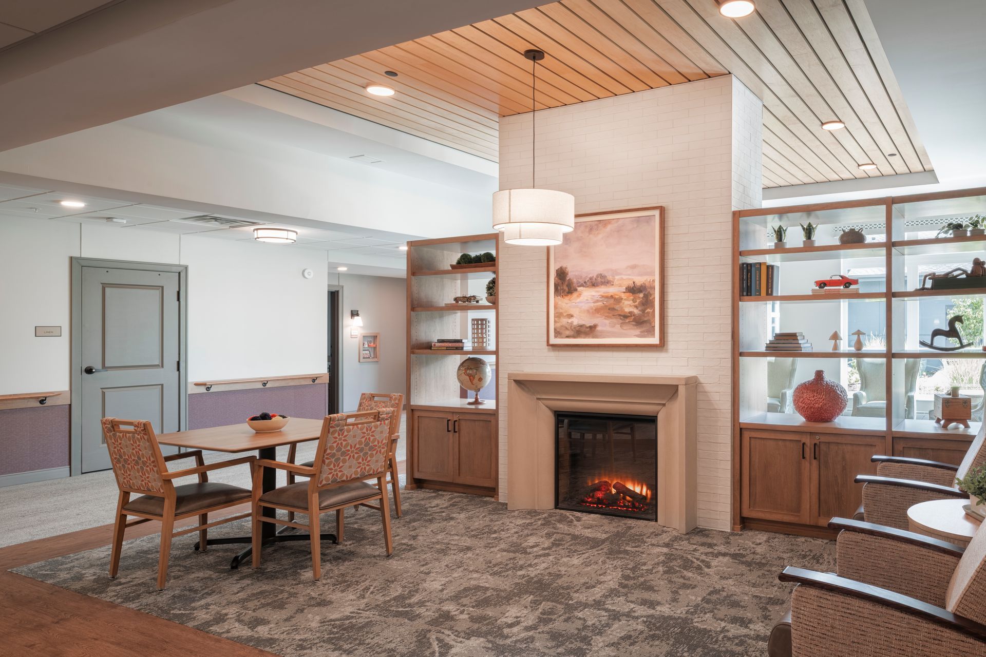 A room with a fireplace, shelving, and a small dining table with chairs, under a slatted wooden ceiling.