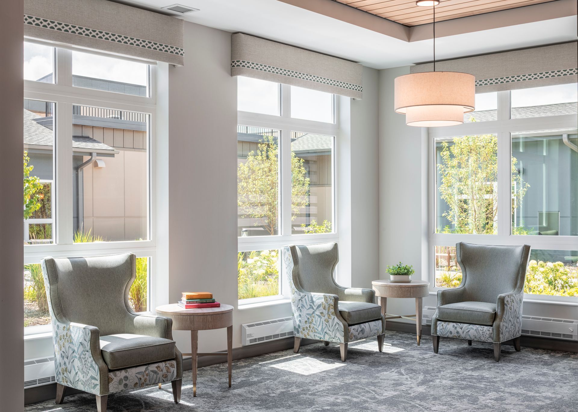 A sunlit room with three grey upholstered armchairs, small side tables, patterned valances, and a modern pendant light.