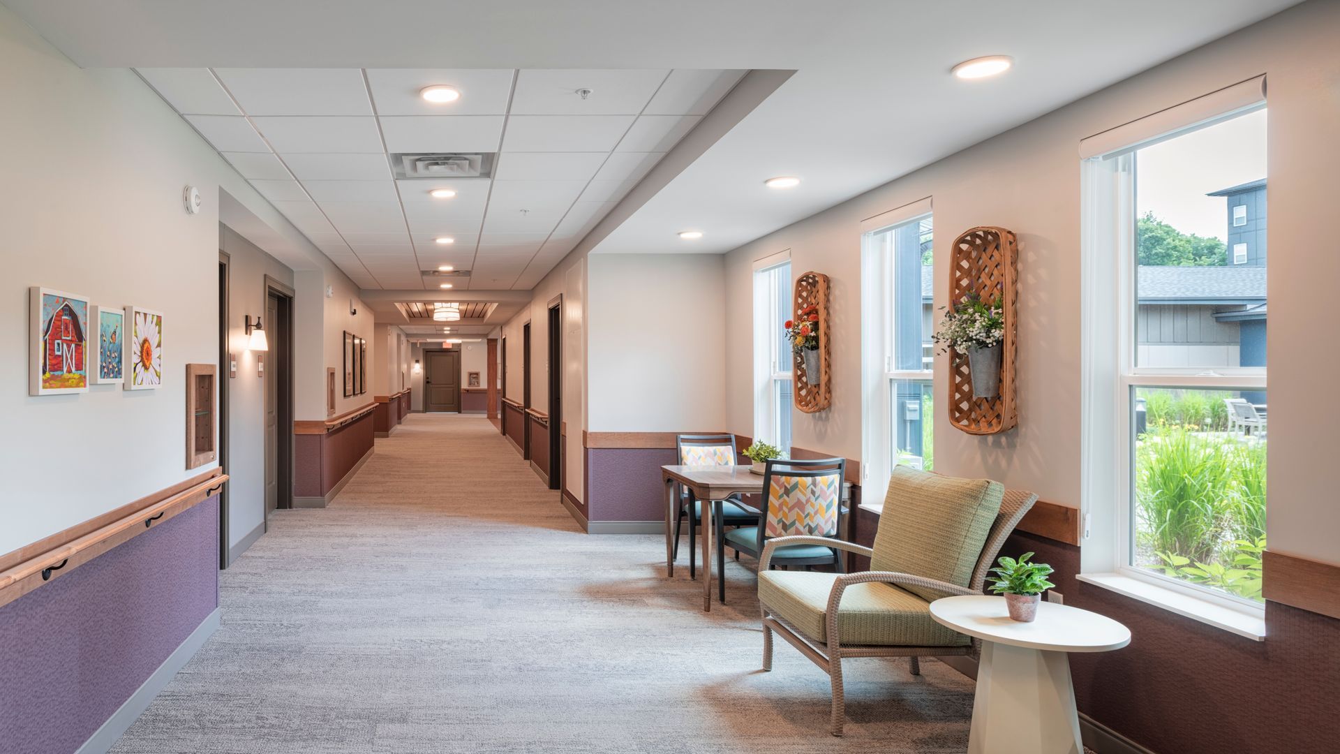 A bright, carpeted hallway with wall art, a small table with chairs, and a seating area by large windows.