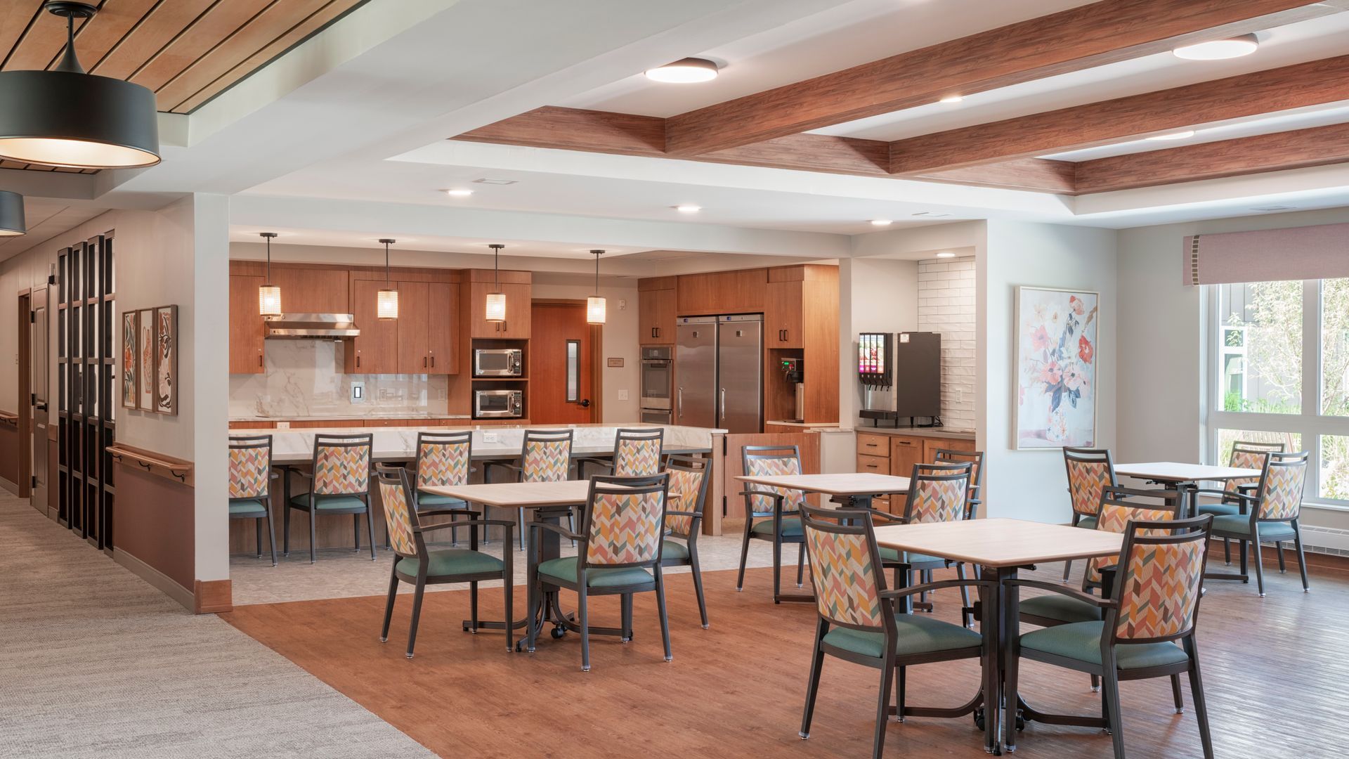 A dining area with square tables, patterned chairs, and a kitchen featuring wooden cabinets and stainless appliances.