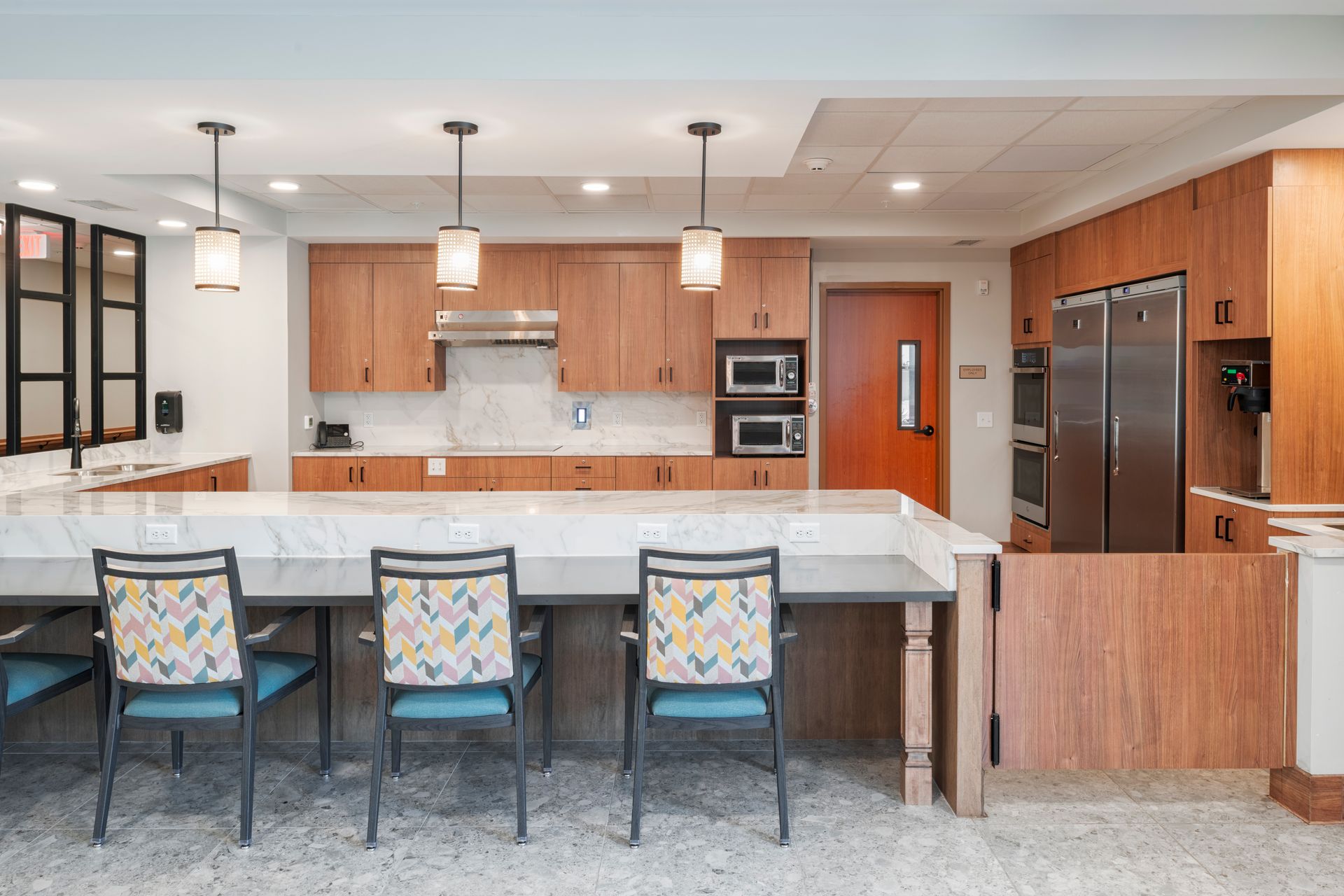 A modern kitchen with wooden cabinets, stainless appliances, and a marble-topped counter with three patterned chairs.