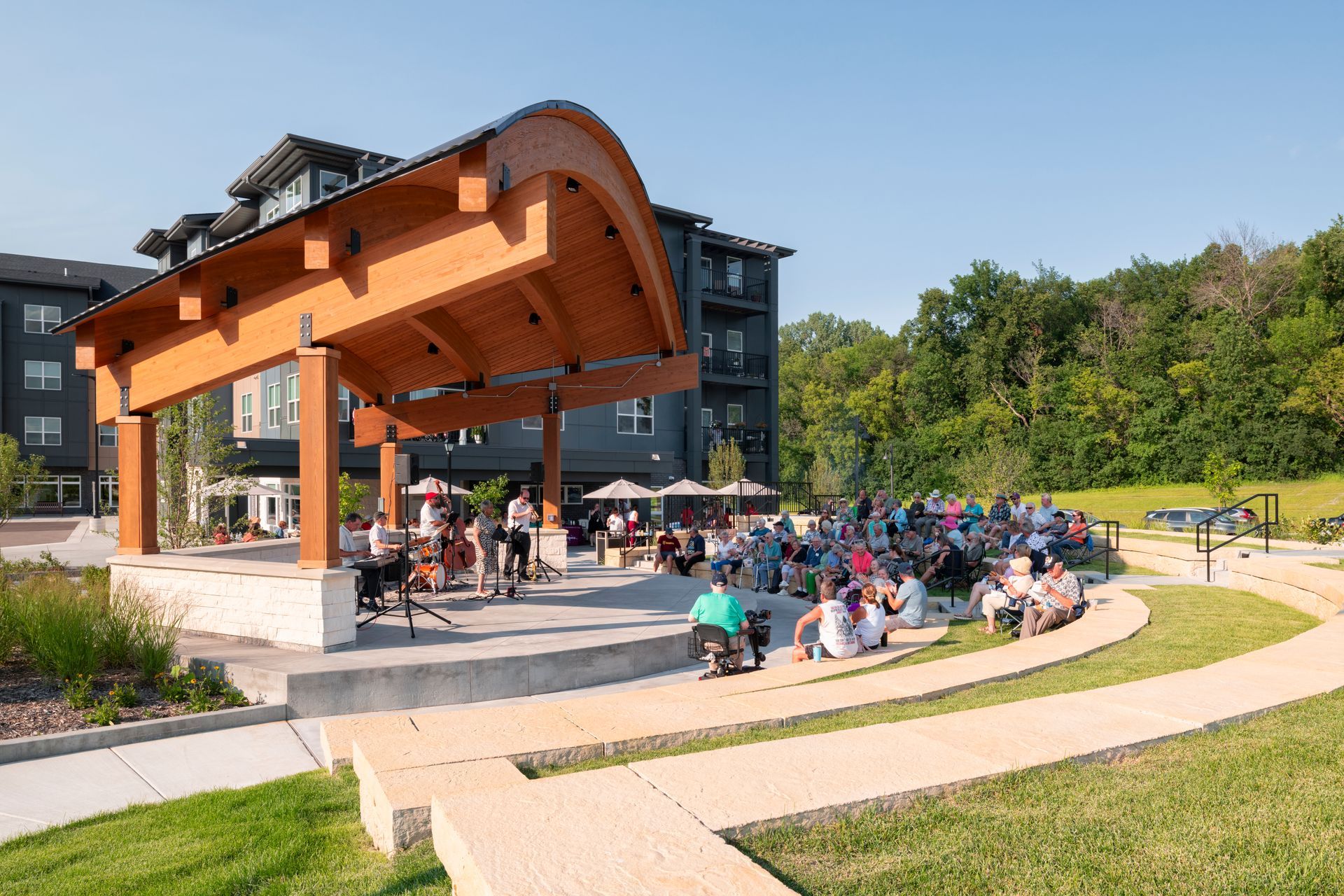 A live band performs under a wooden pavilion for an audience seated on grass and terraced stone seating at an outdoor park.
