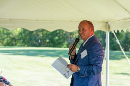 A man in a blue suit holds a microphone and papers while speaking under a white canopy outdoors.