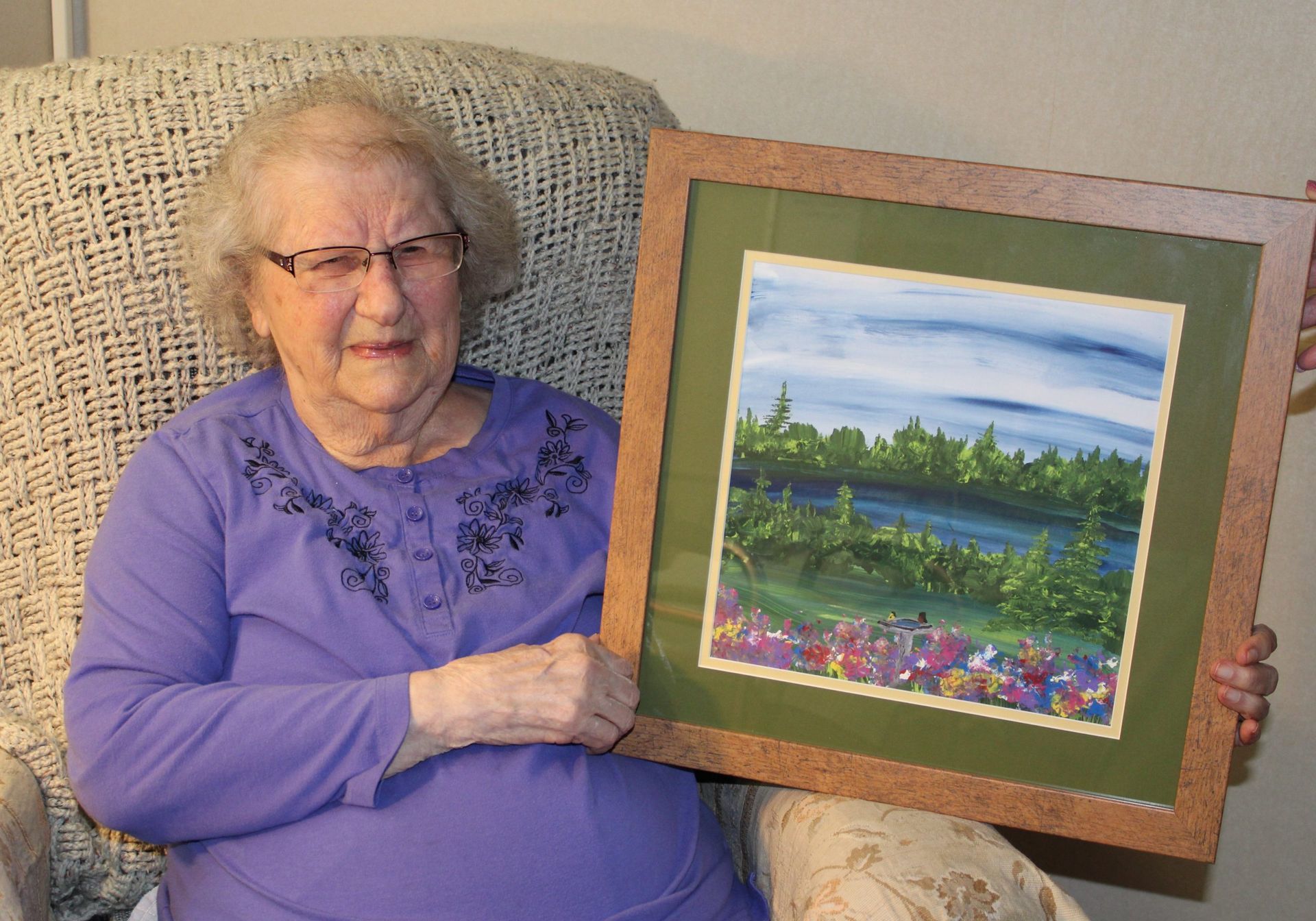 A seated person holds a framed landscape painting featuring a forest, a blue lake, and a field of colorful wildflowers.