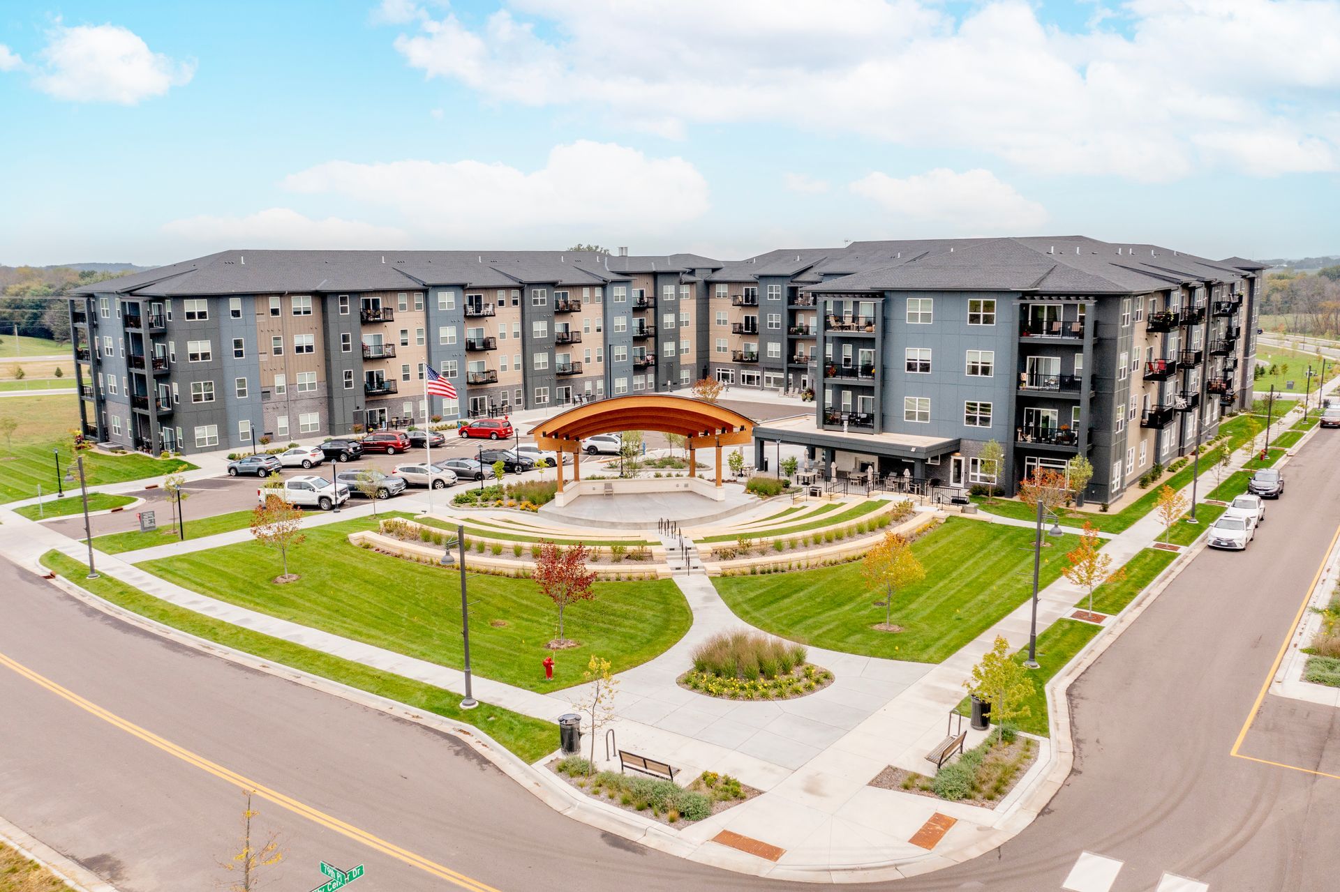 A modern multi-story apartment building with dark grey siding, balconies, and a paved entrance with outdoor seating.