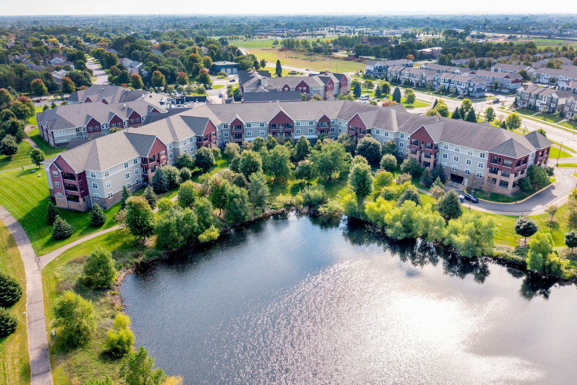 Aerial view of an apartment complex with tan and red brick siding, surrounded by trees and a pond in a suburban setting.