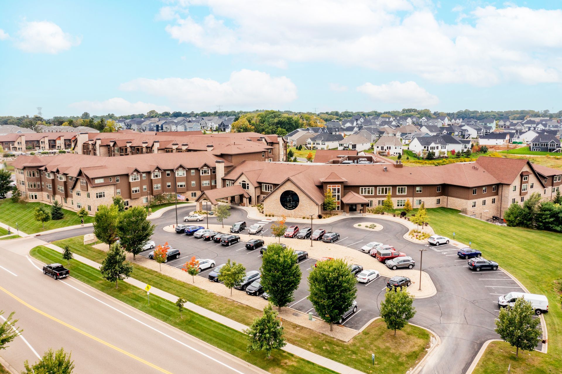 Aerial view of a tan-roofed apartment complex with parking lot and road in a suburban neighborhood