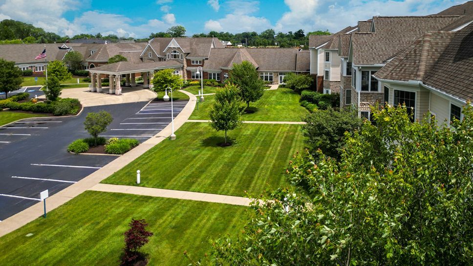 A large multi-story suburban building with a parking lot and a manicured green lawn under a partly cloudy sky.