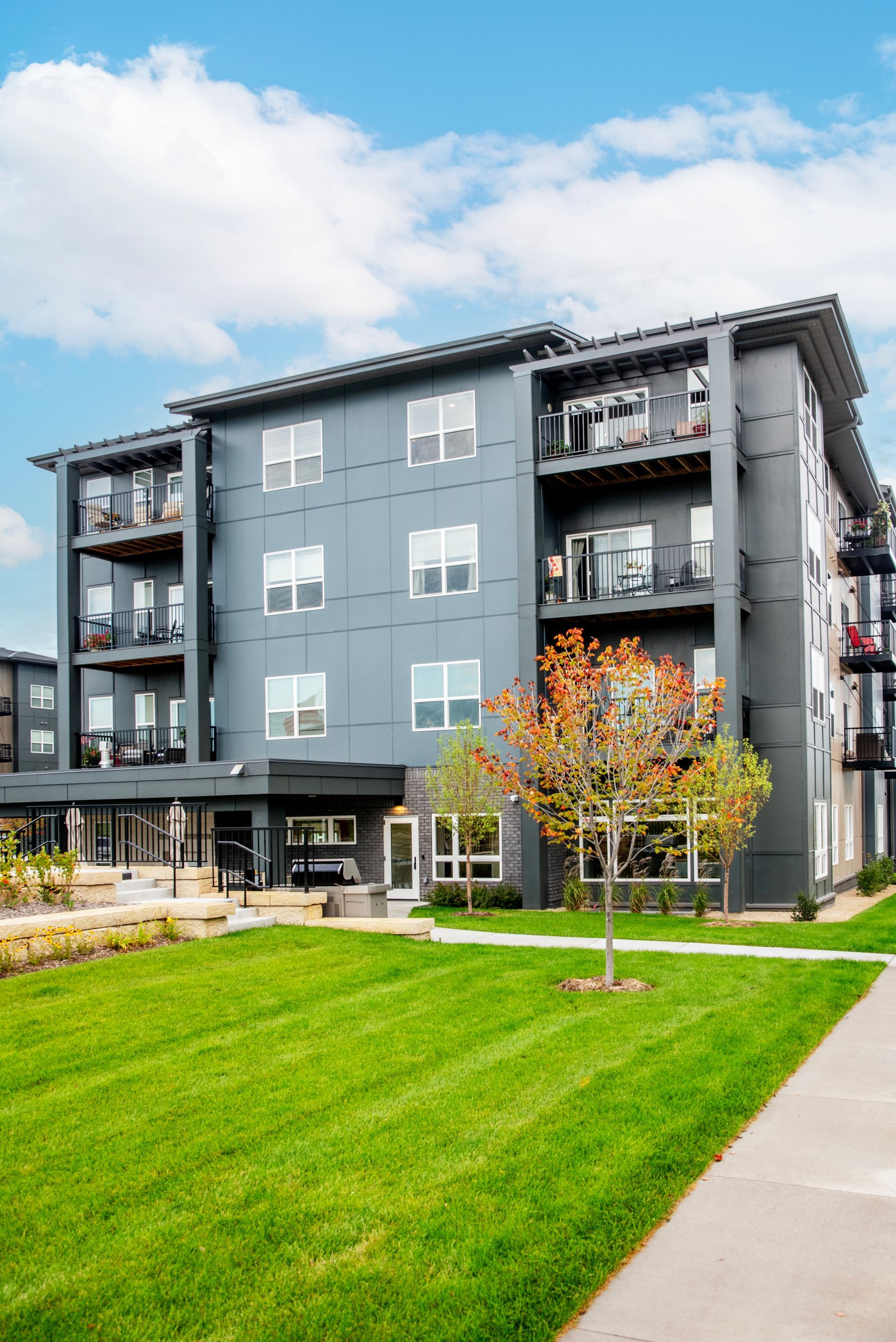 A modern, dark-gray four-story apartment building with balconies, surrounded by a lawn and a tree under a blue sky.