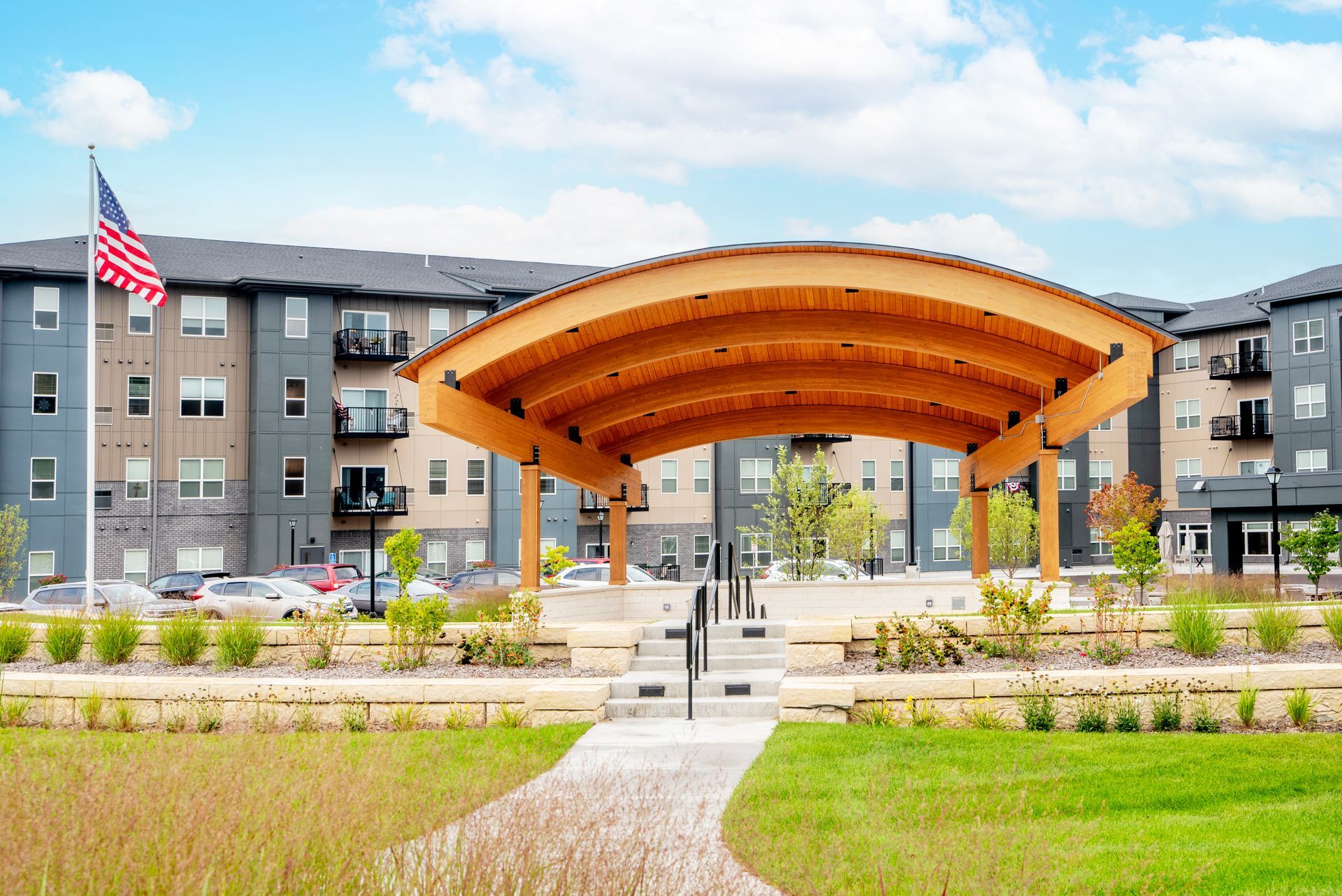 A large wooden arch pavilion stands in front of a modern multi-story apartment complex with an American flag flying.