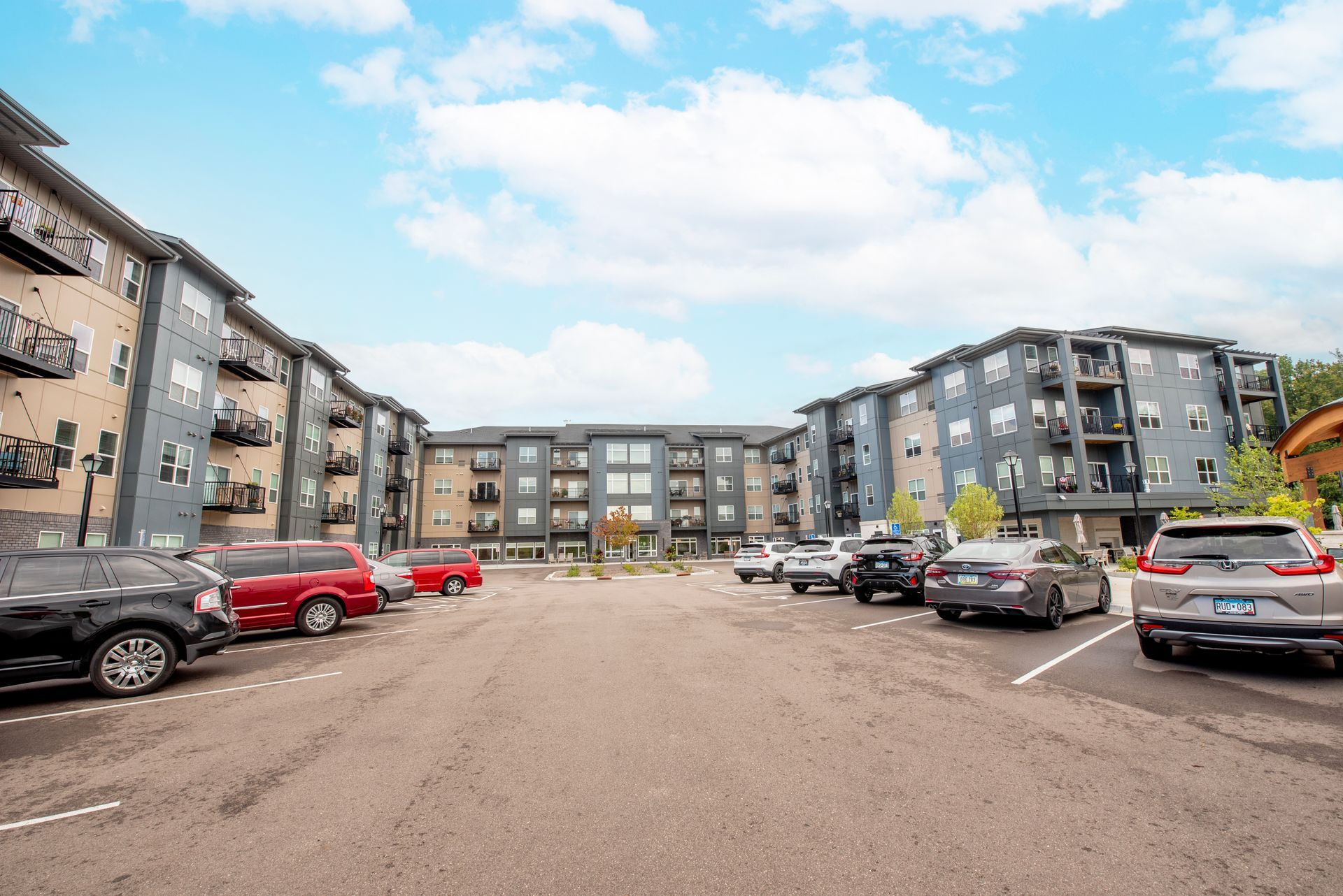 A parking lot surrounded by modern, multi-story apartment buildings under a partly cloudy sky.