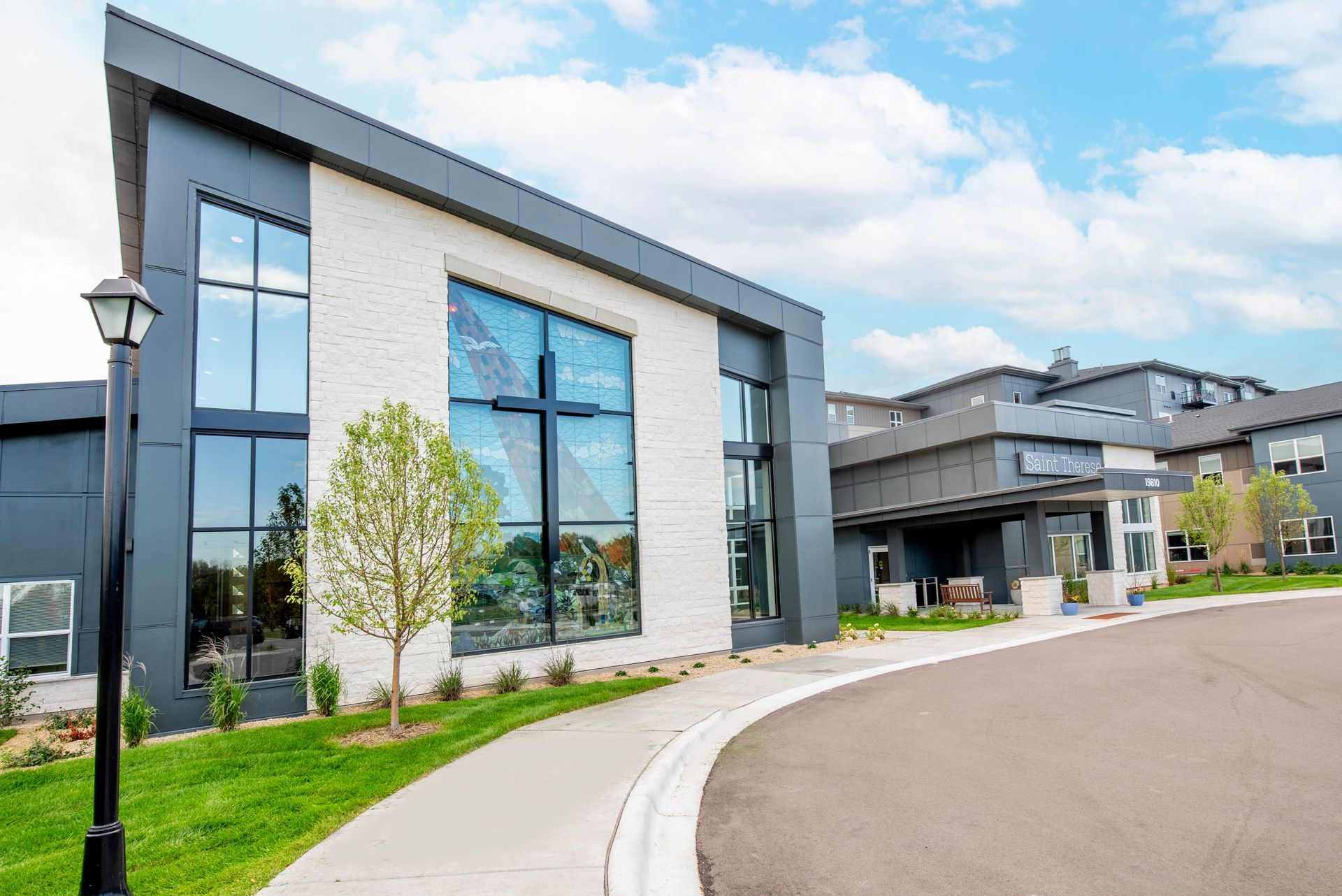 Modern building exterior with white stone, dark panels, large windows featuring a cross, and a curved driveway.