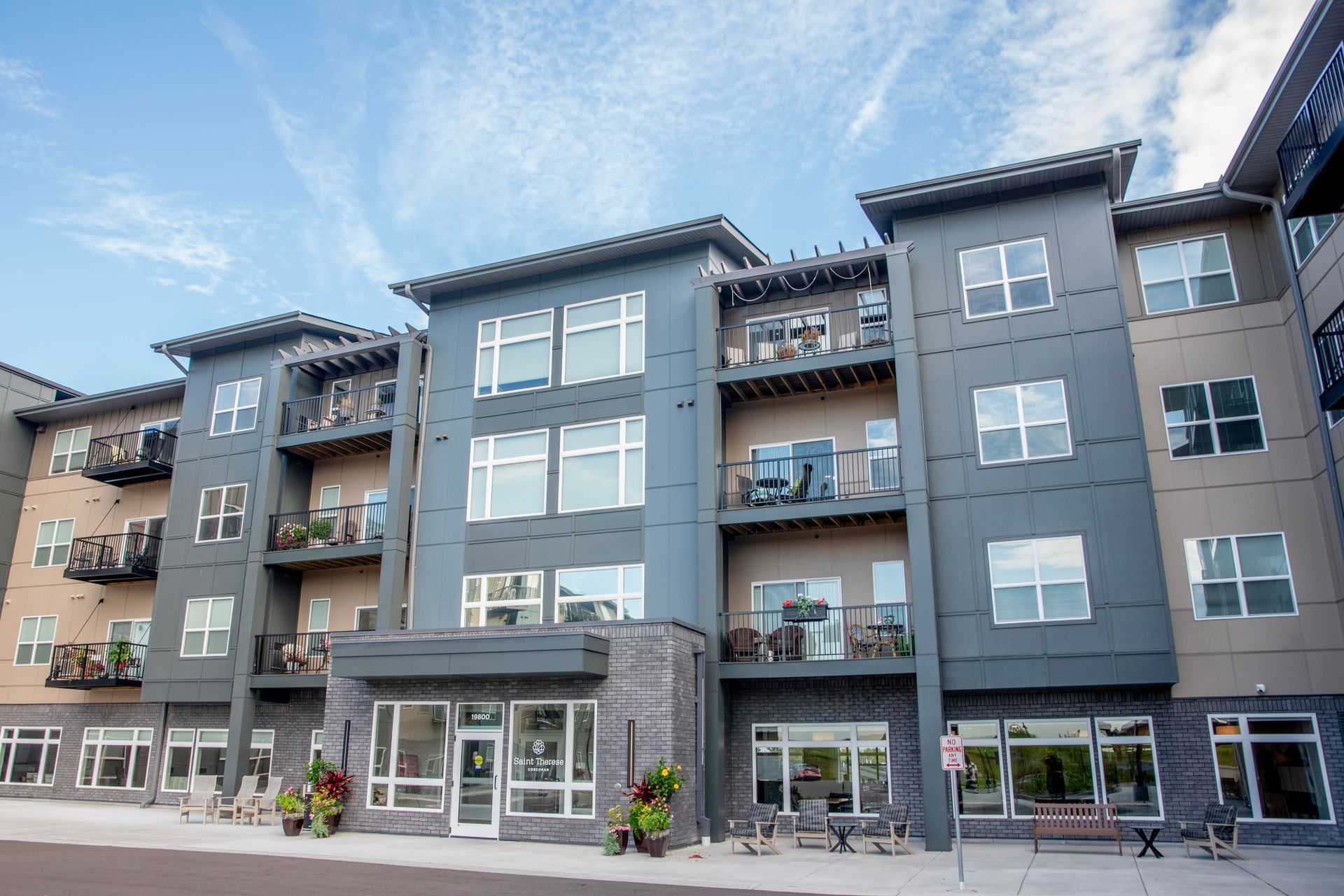 A multi-story modern apartment building featuring gray and beige siding, balconies, and a ground-floor entrance.