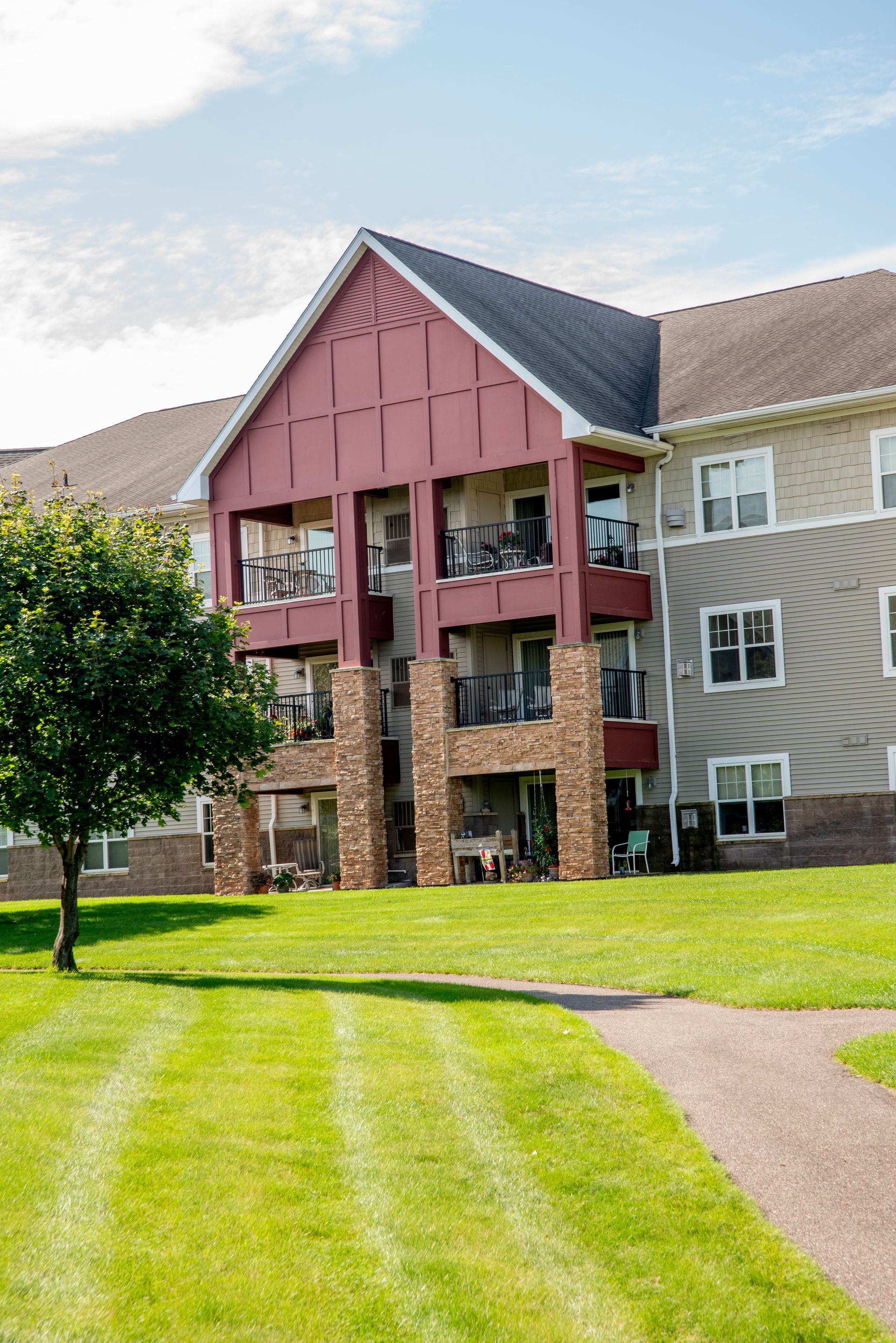 A multi-story apartment building with a red-accented facade, stone pillars, and balconies, set beside a manicured lawn.