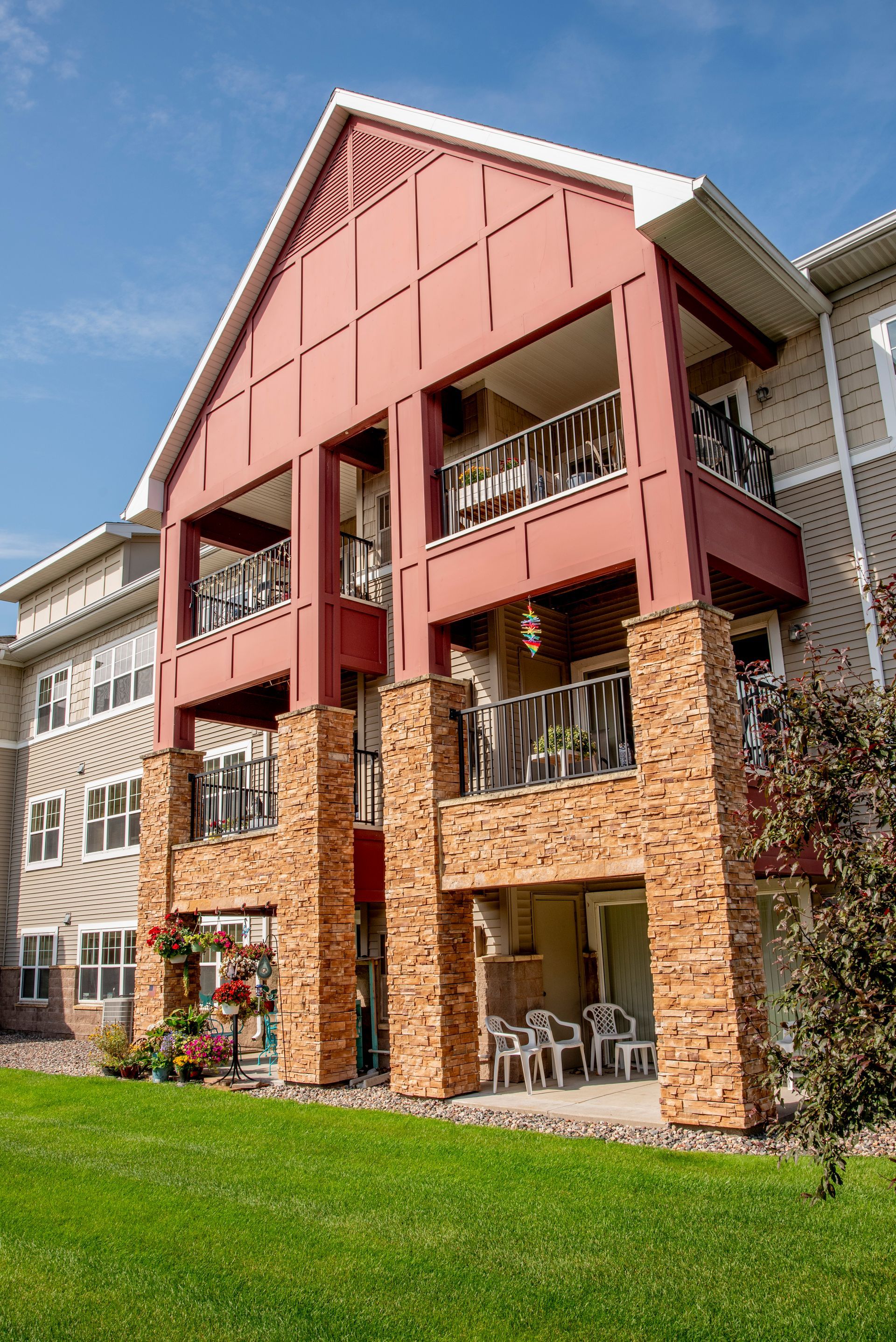 Three-story apartment building with a reddish-brown facade, stone pillars, and balconies, set against a grassy lawn.