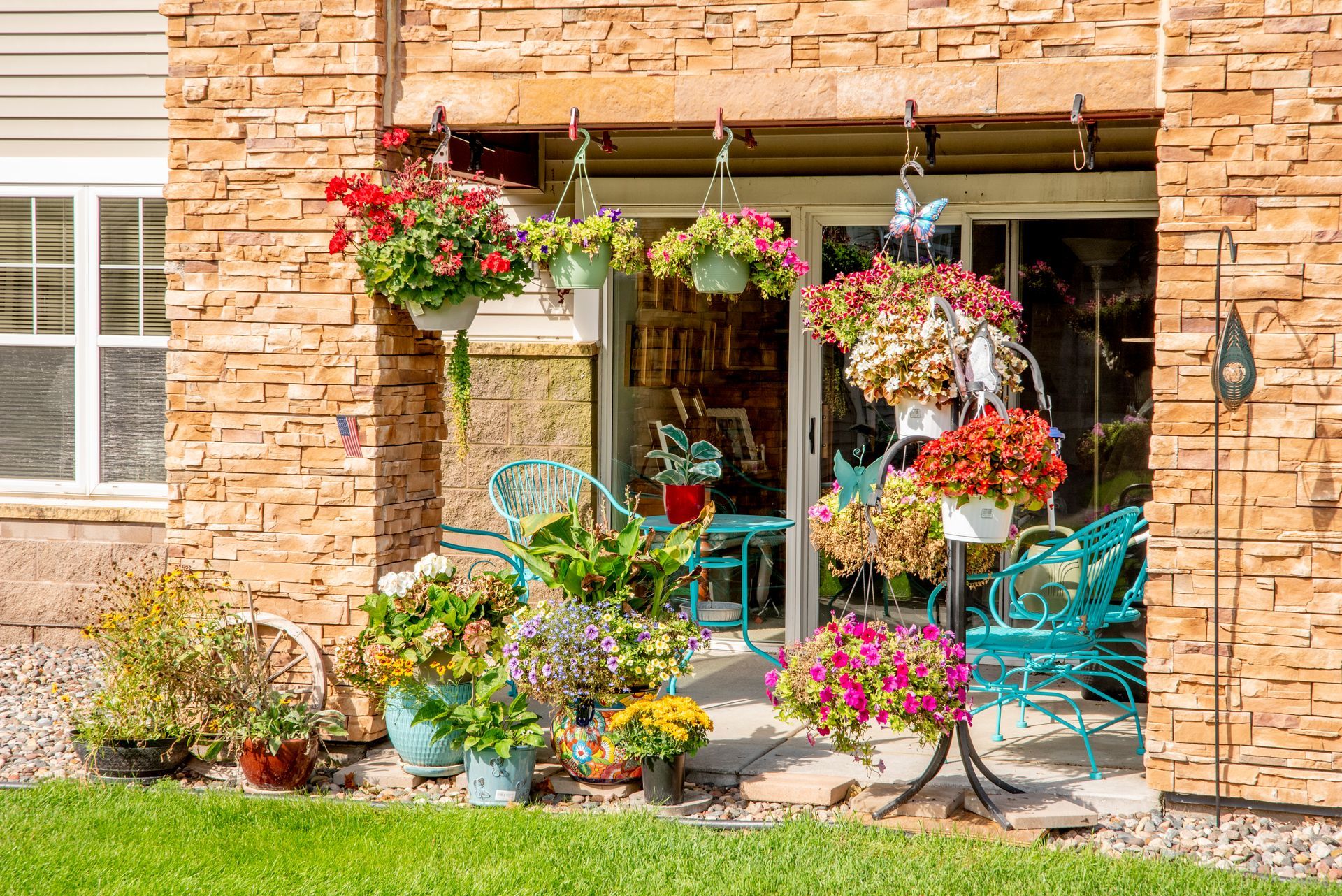 A patio with many hanging and potted flower arrangements, along with turquoise metal chairs, against a stone wall.