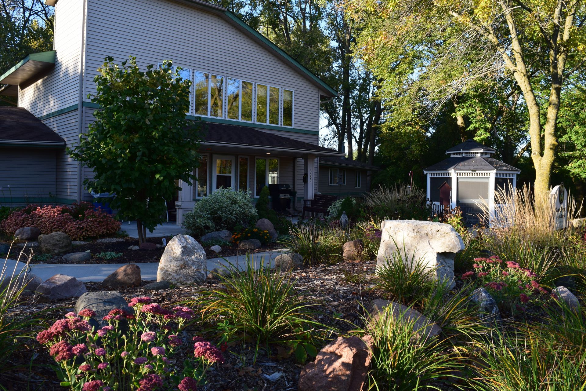 A multi-level home with a large garden, rock landscaping, and a gazebo in the backyard on a sunny day.