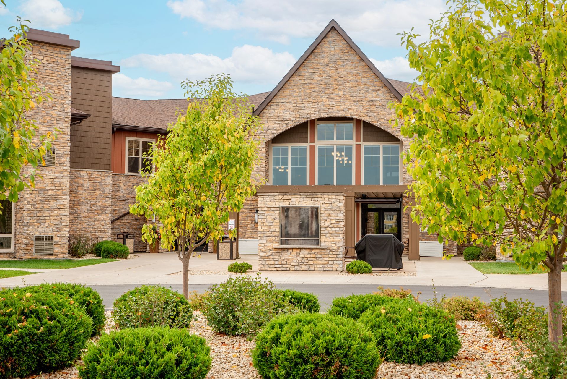 Stone building entrance with arched windows, flanked by green trees and landscaped shrubs.
