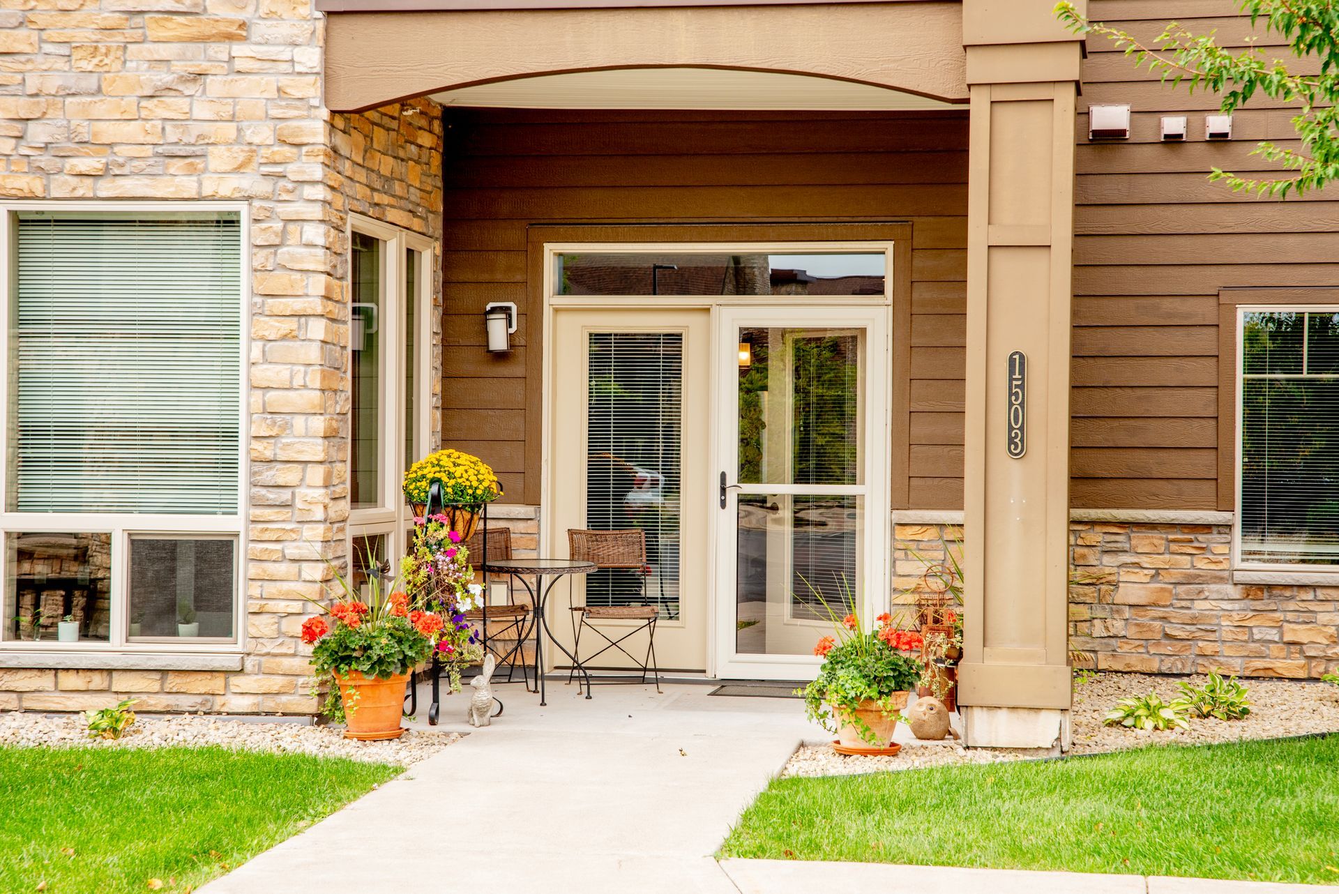 Apartment entrance with stone facade, glass doors, and potted flowers beside a walkway