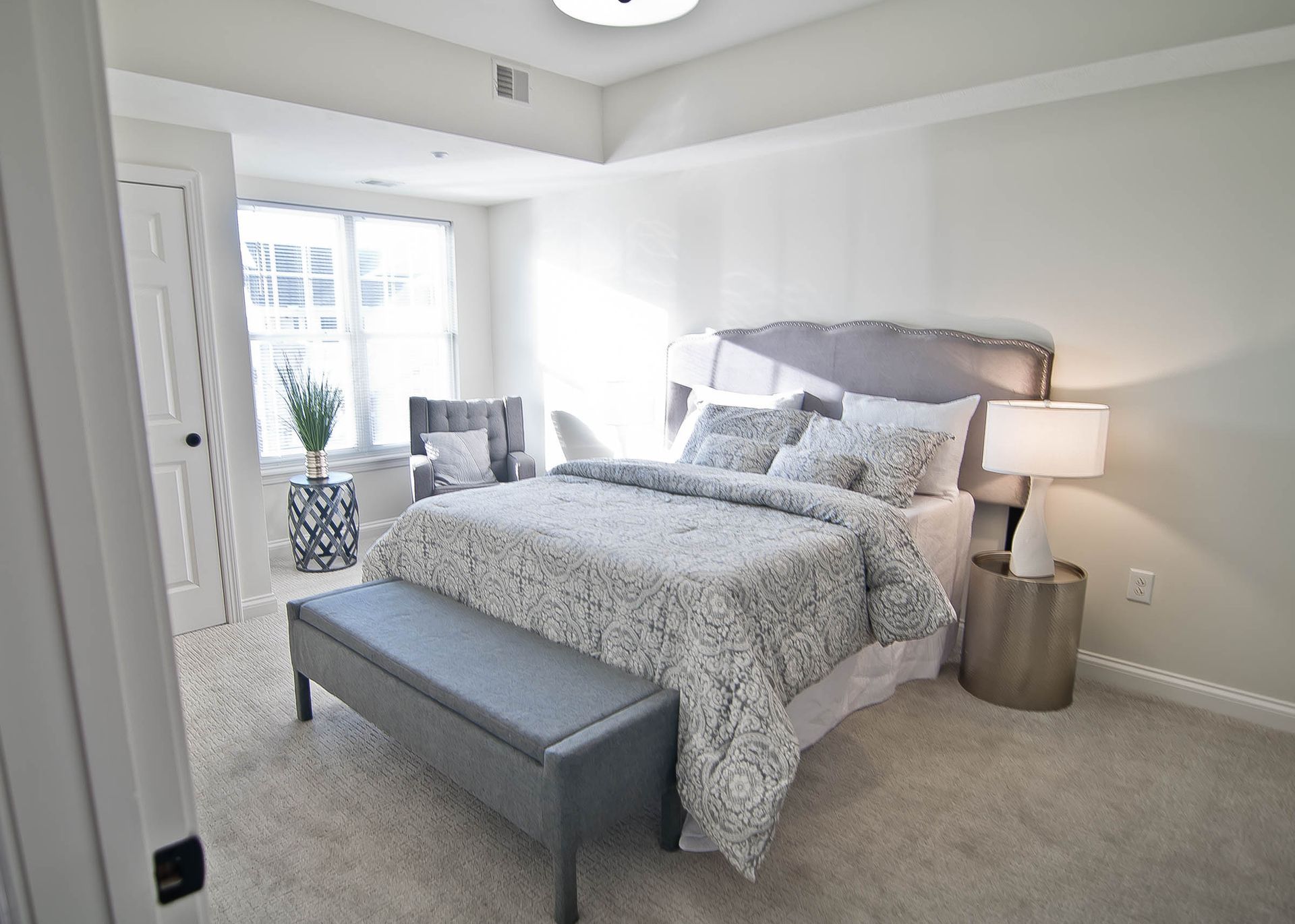 A bedroom with a grey tufted bed, patterned comforter, matching bench, bedside table with a lamp, and a window with a chair.
