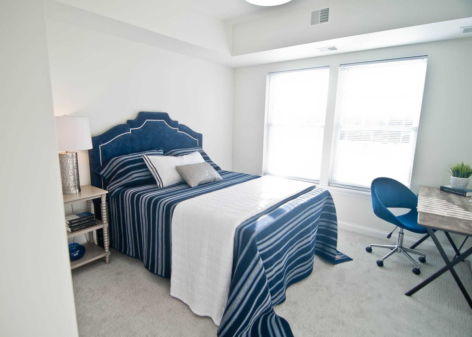 A bedroom with a blue-and-white striped bedspread, a blue upholstered headboard, a bedside lamp, and a desk with a chair.