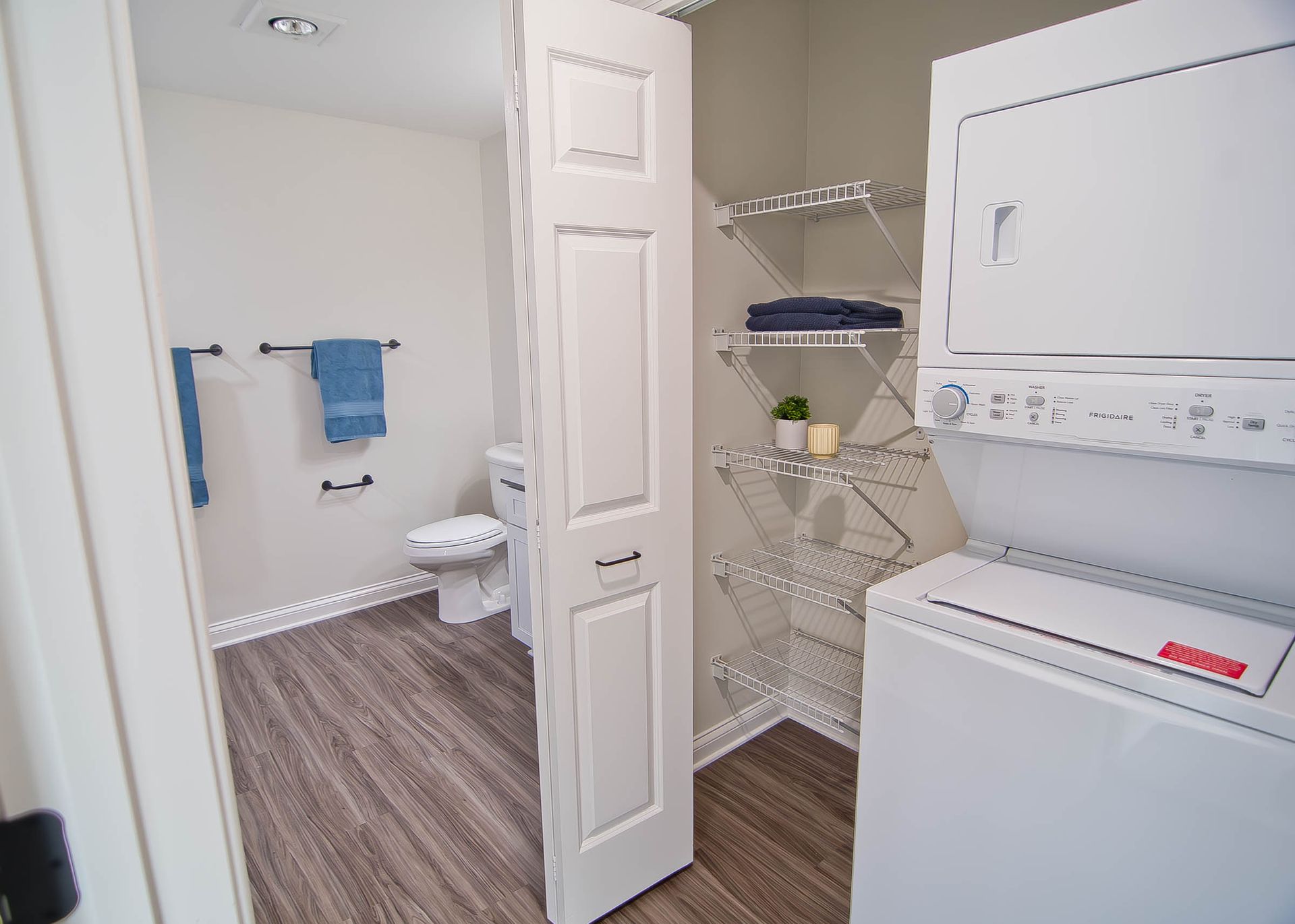 A laundry room with a stacked washer and dryer, open white shelving, and a doorway leading to a bathroom.
