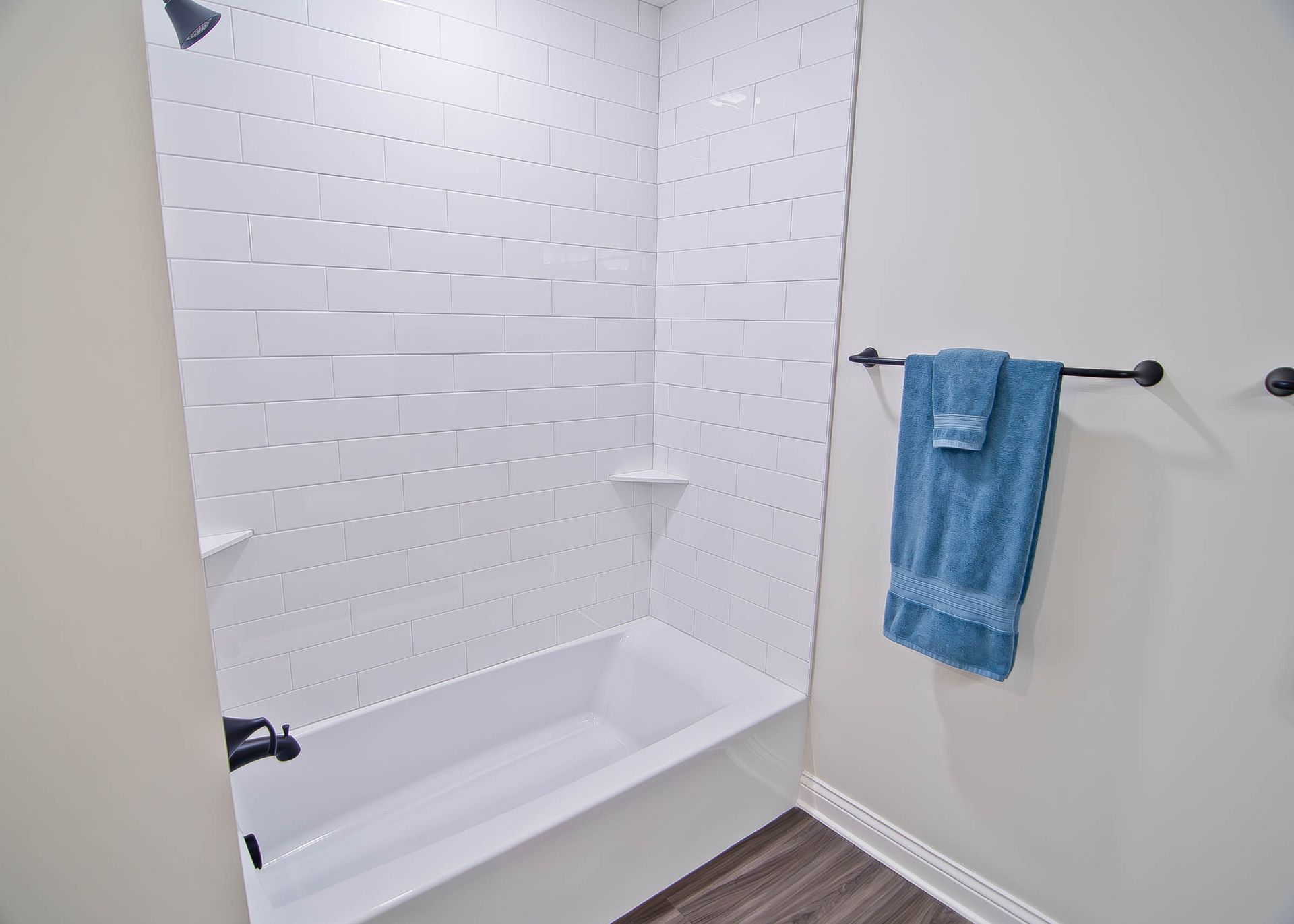 A white bathtub with a tiled surround and a black shower fixture next to a towel hanging on a dark rod against a wall.