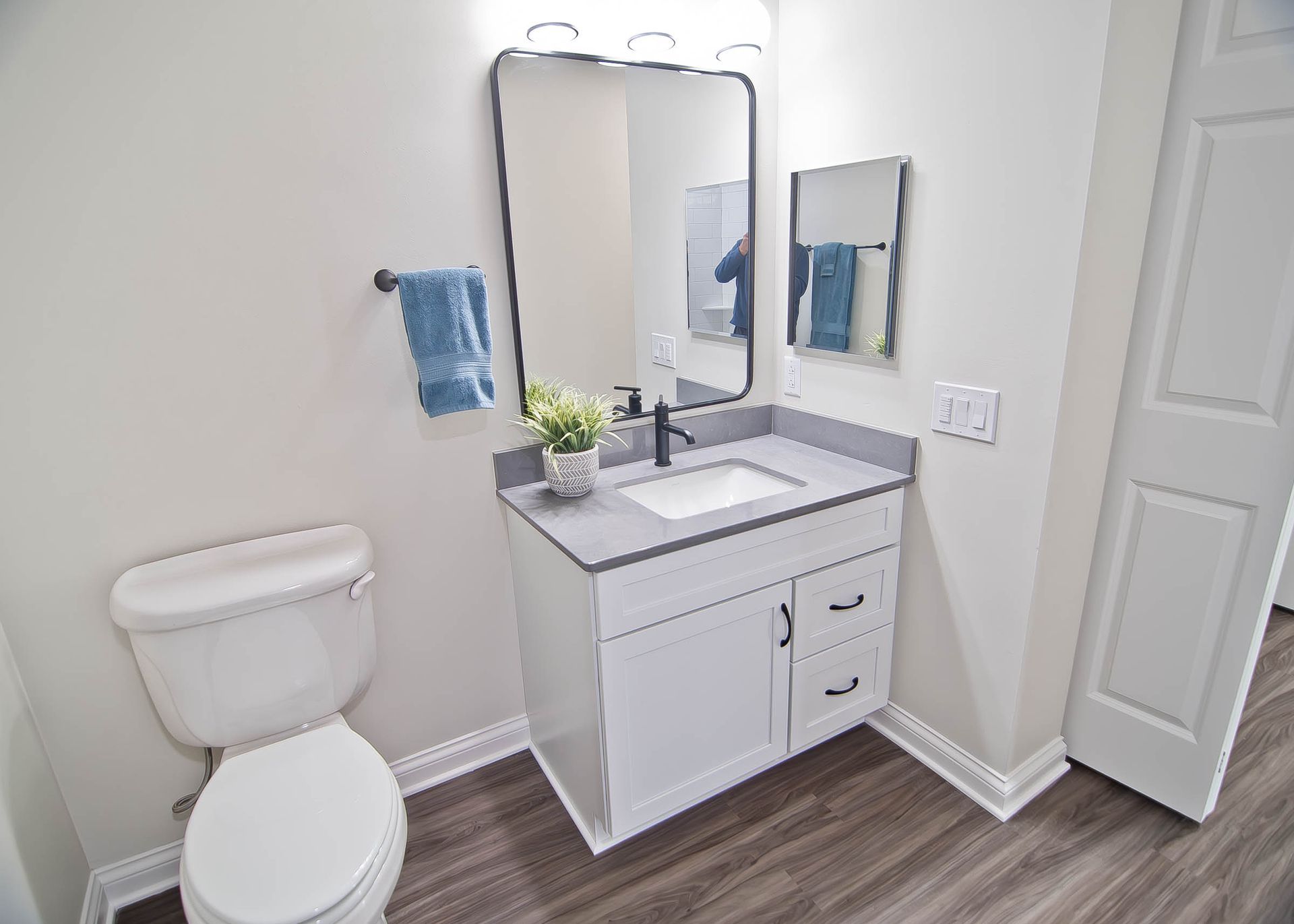 A modern bathroom with a white vanity, grey countertop, sink, mirror, toilet, and blue towel on a grey wood-look floor.