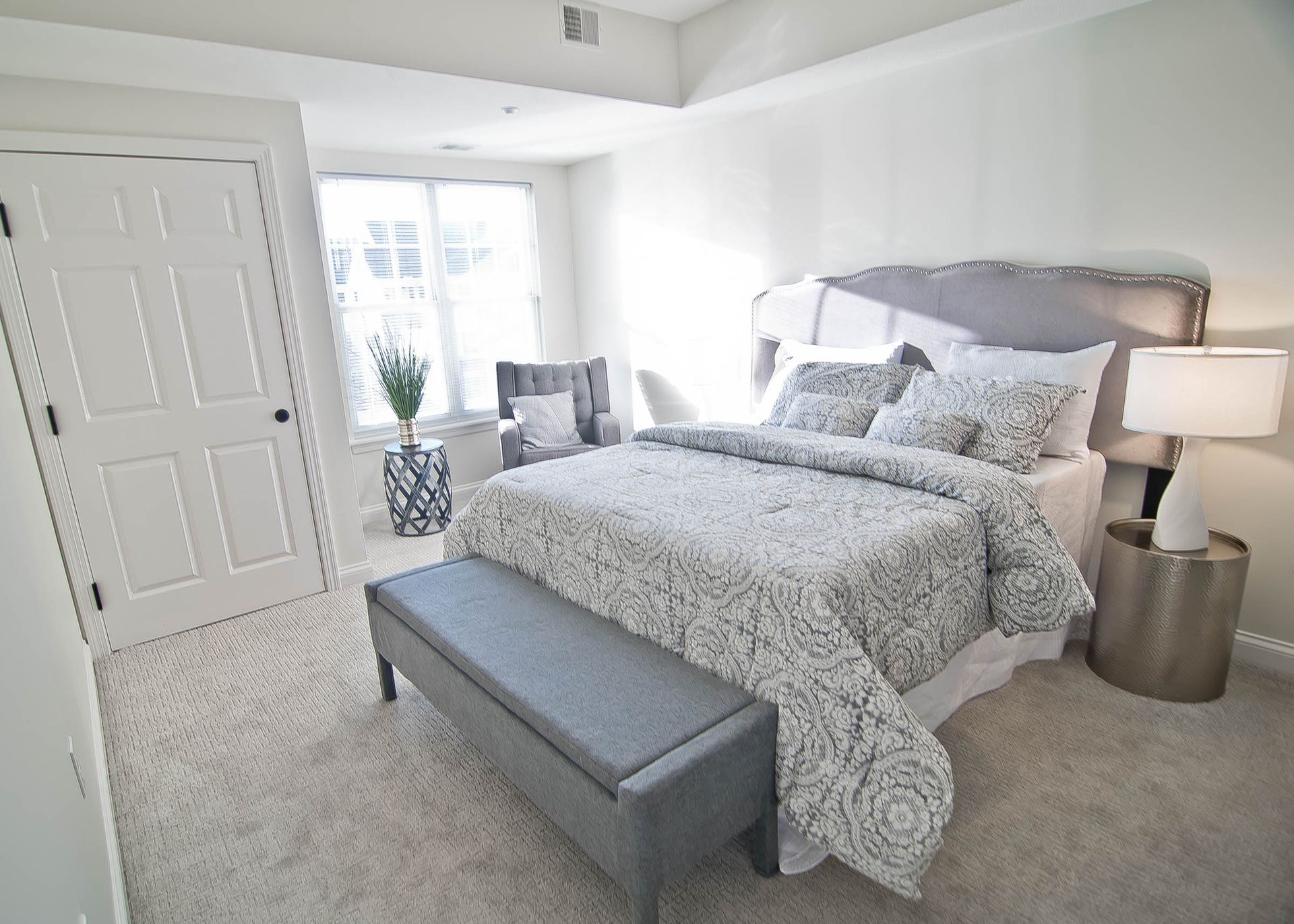 A bright bedroom featuring a queen bed with a grey patterned comforter, a grey bench, and a modern silver bedside lamp.