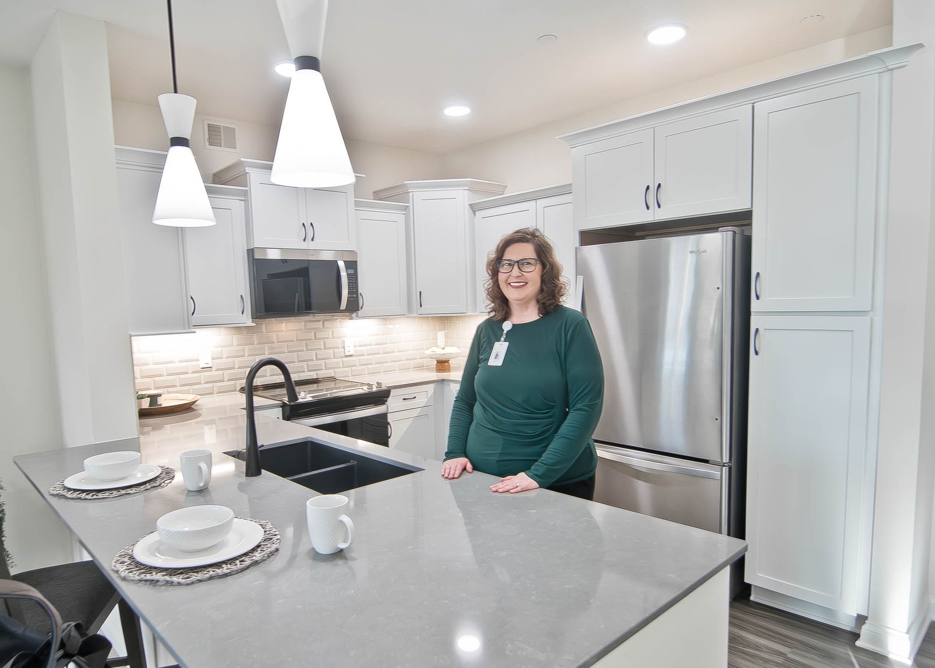 Woman standing in a bright white kitchen beside a stainless steel refrigerator and marble island.
