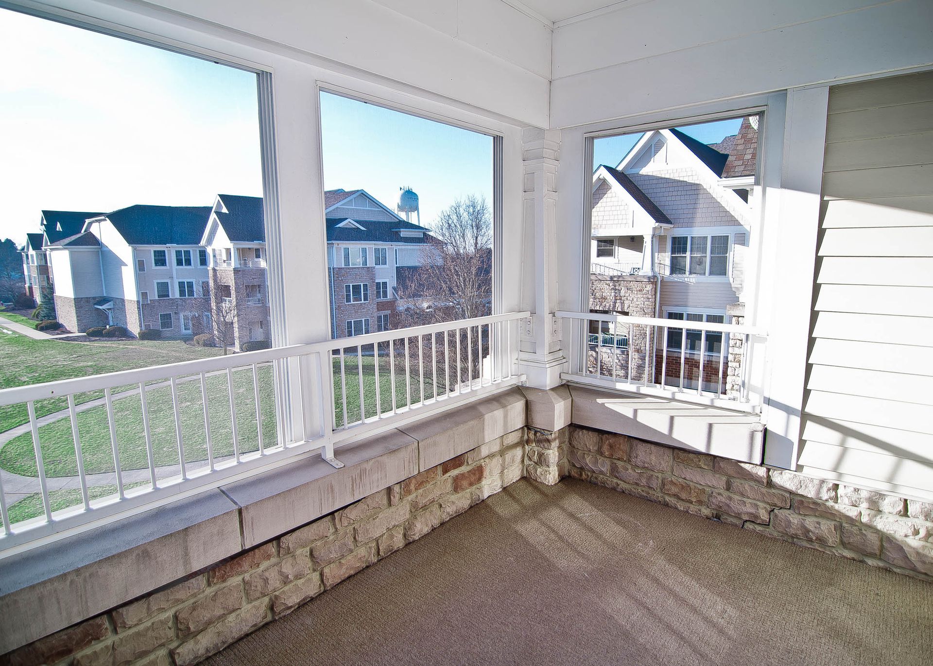 A sunlit, covered stone balcony with white railings overlooks a residential area with apartment buildings.