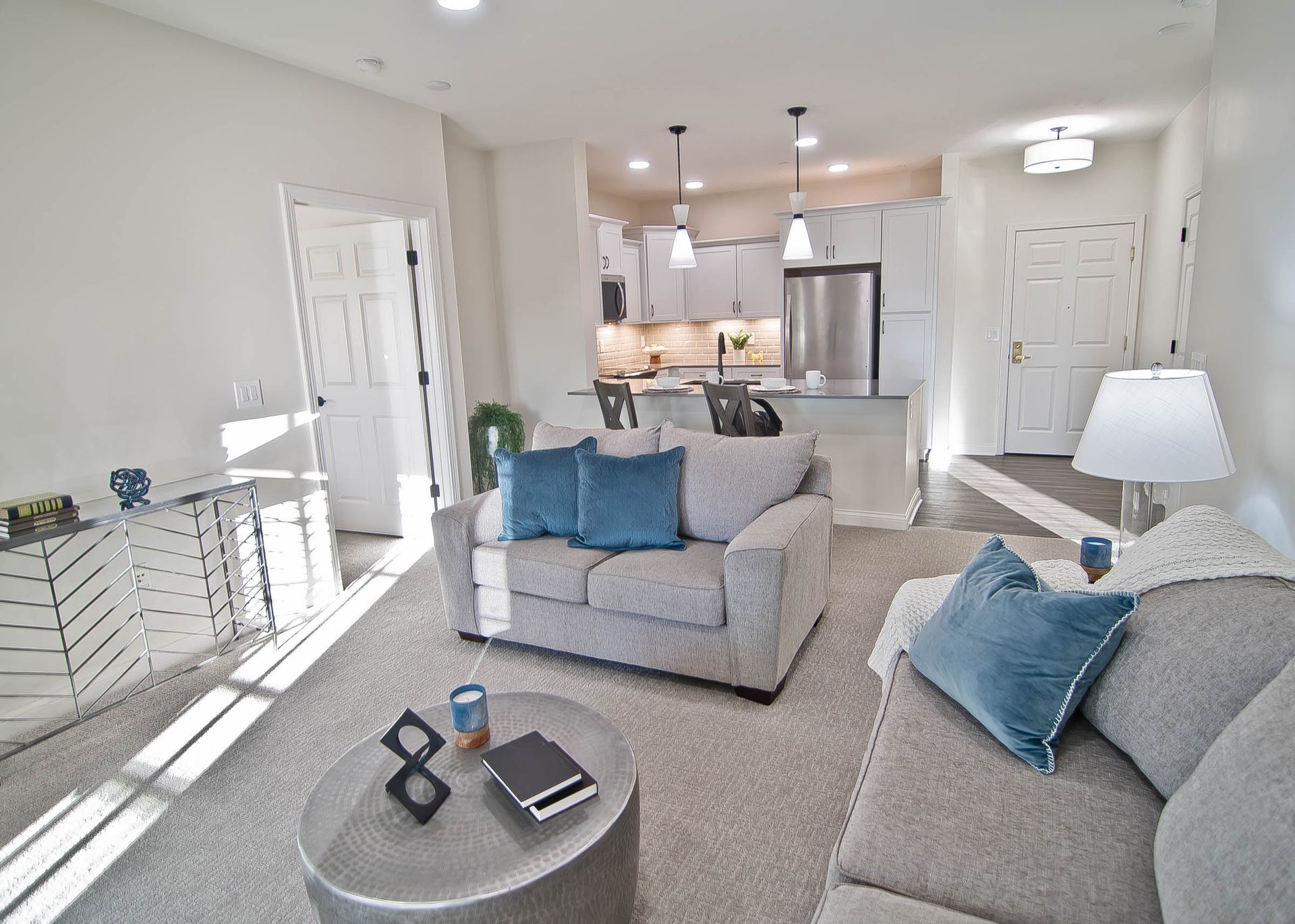 A bright, modern living room featuring a grey couch with blue pillows, a circular coffee table, and an open kitchen.