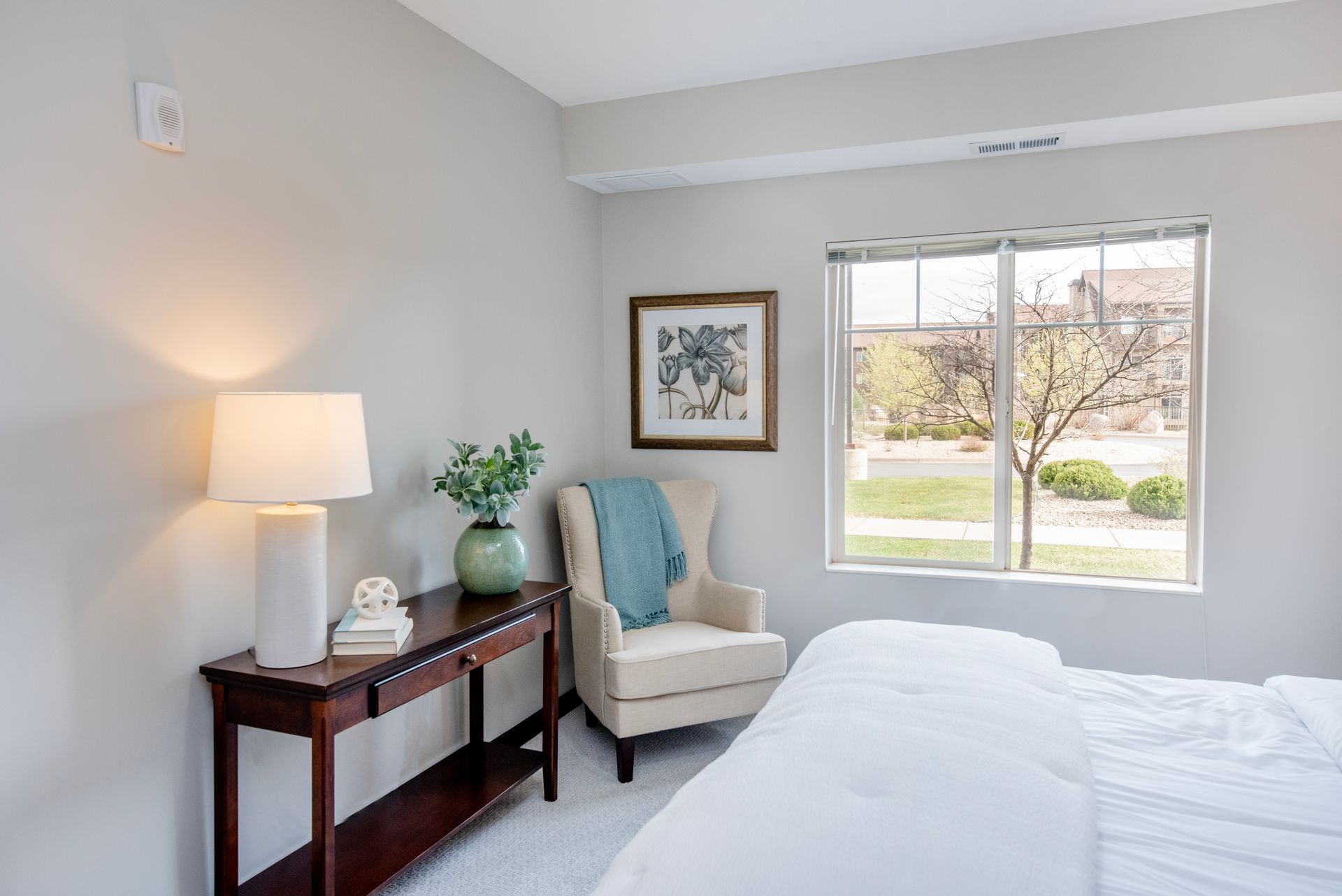 Bright bedroom with bed, armchair, lamp, console table, and window view of trees