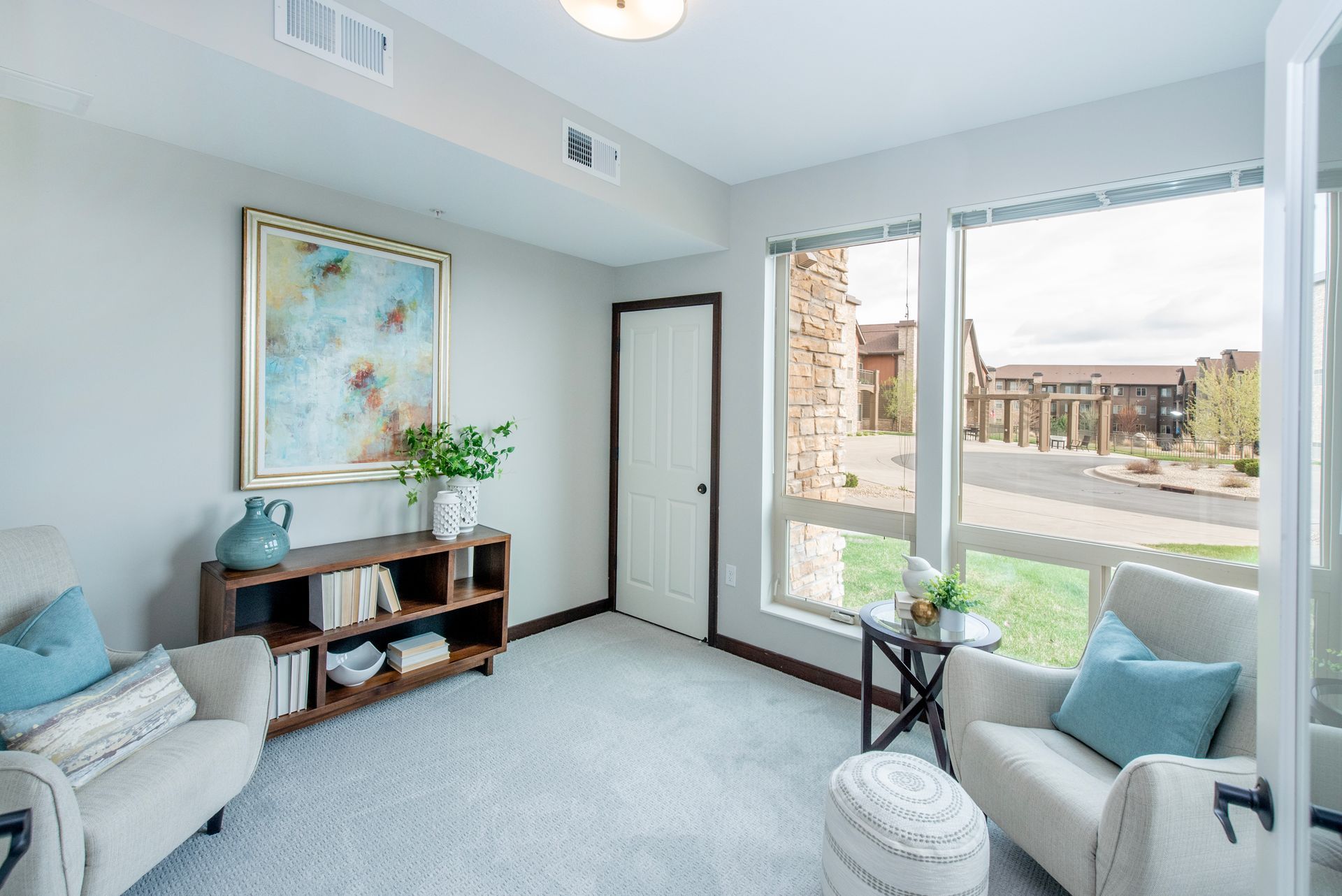 Bright living room with light blue walls, two armchairs, a side table, and large windows overlooking a neighborhood.