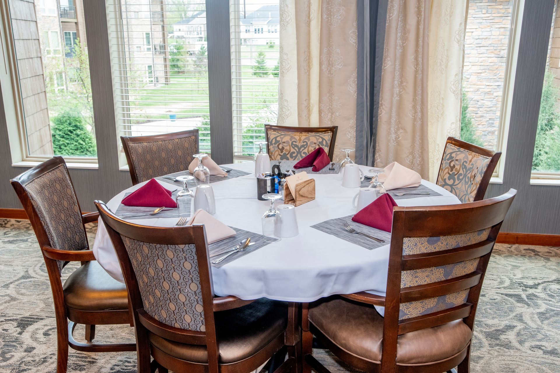 Round dining table set near large windows, with folded napkins and chairs in a bright room.