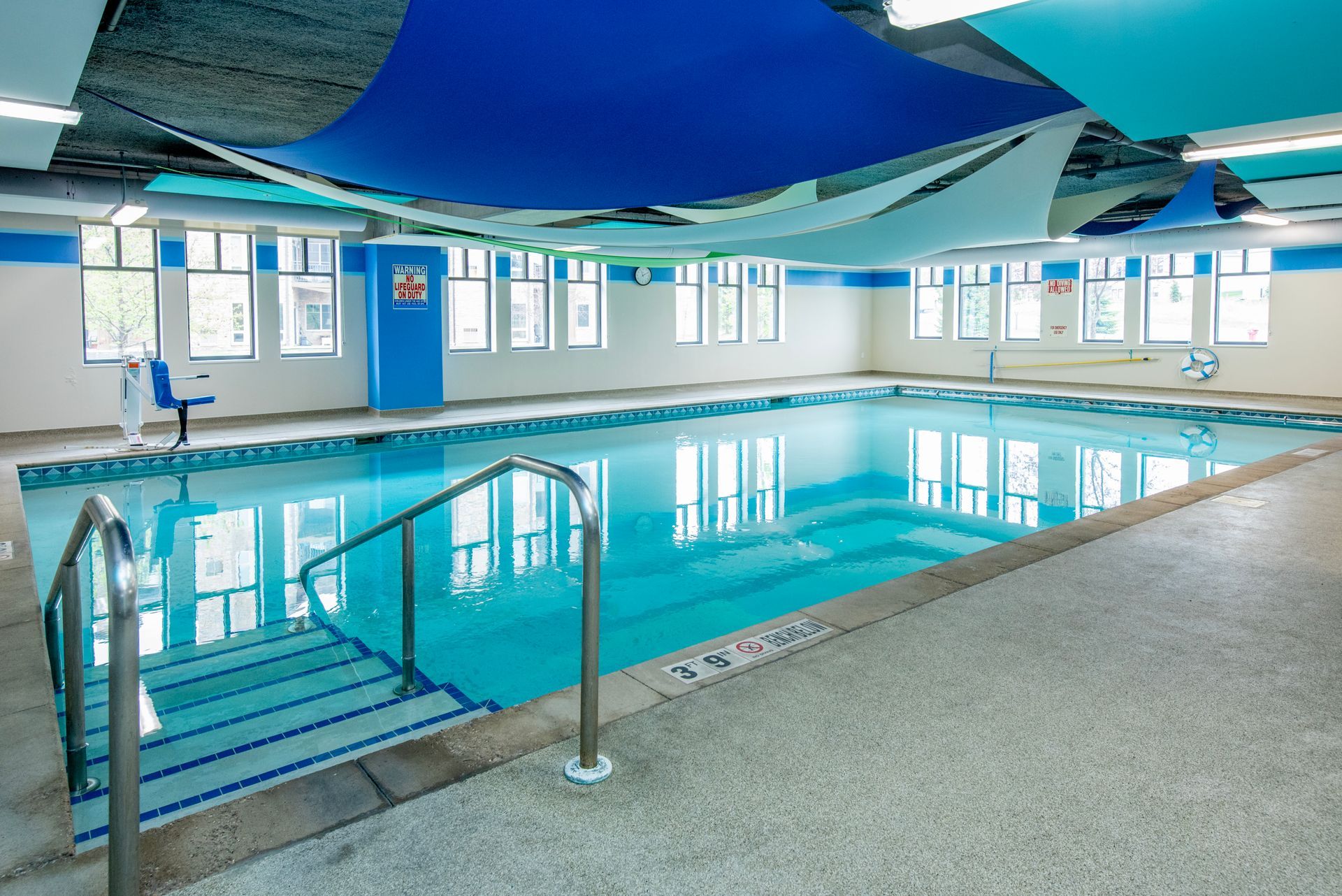 Indoor swimming pool with blue ceiling, white walls, and metal handrails by the steps.