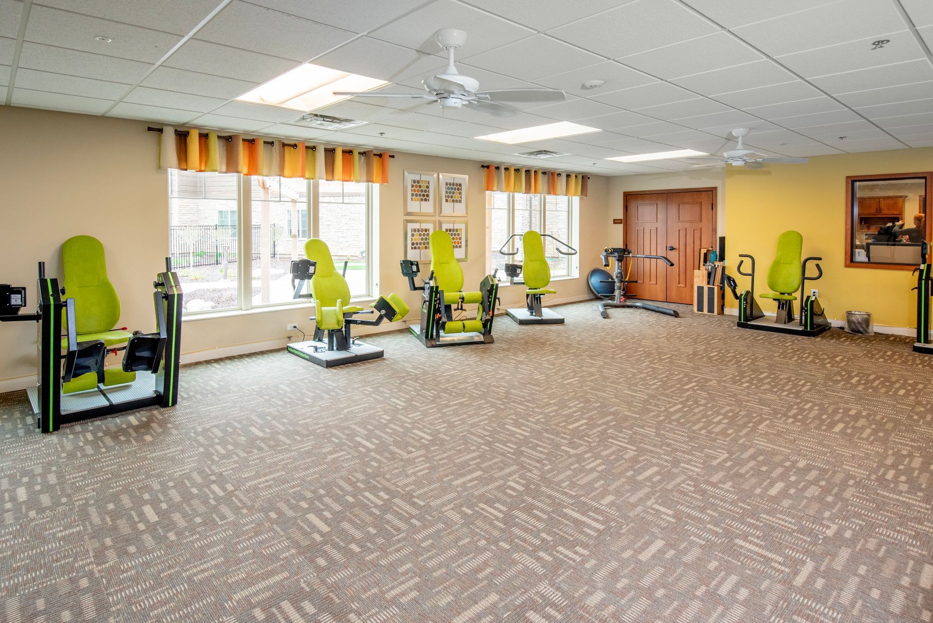 Bright fitness room with green exercise machines, large windows, and patterned carpet