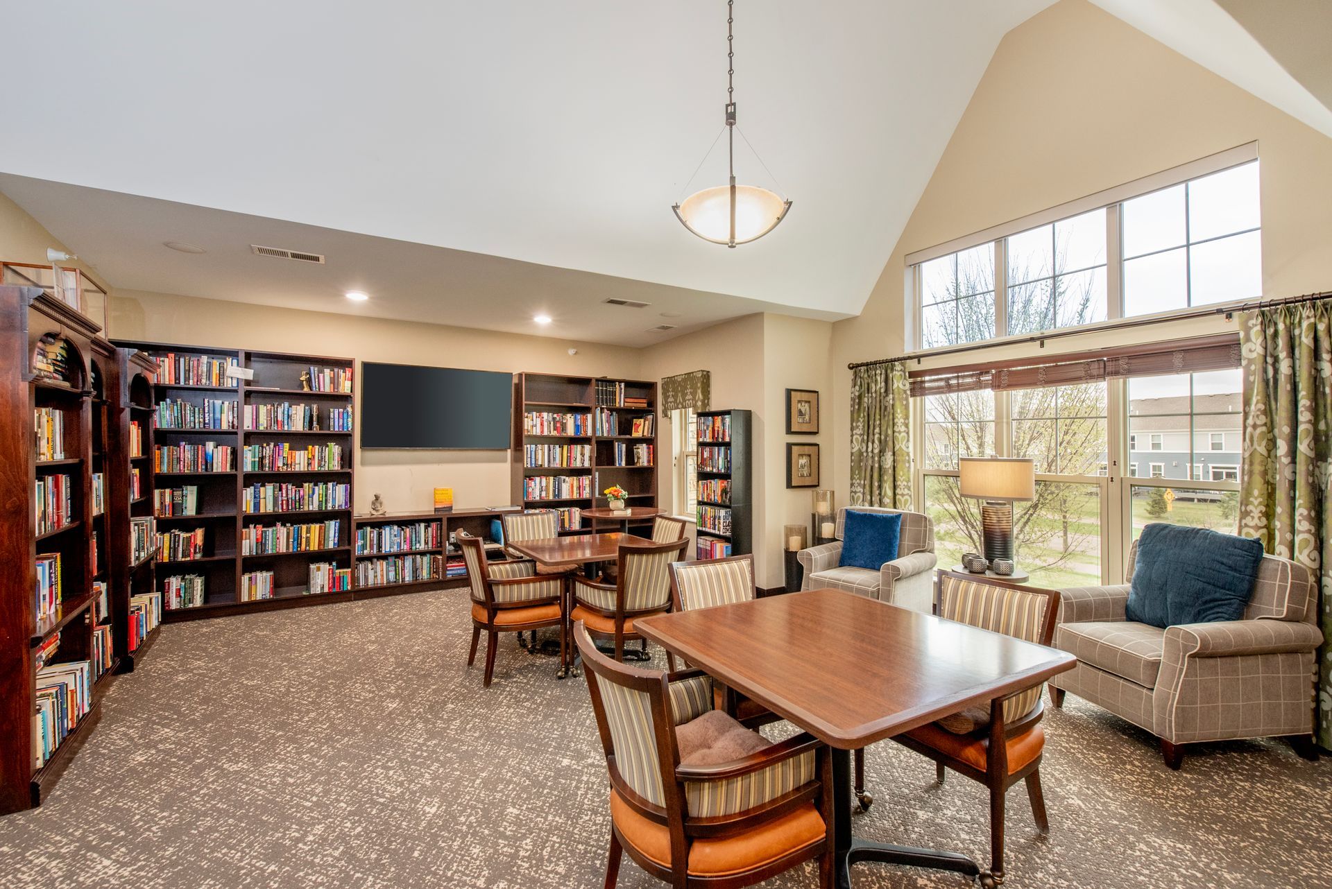 Quiet library lounge with bookshelves, tables, chairs, and a bright seating area by large windows.