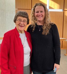 Two smiling people standing side-by-side in front of light wood paneling, one wearing a red blazer, the other in a black top.