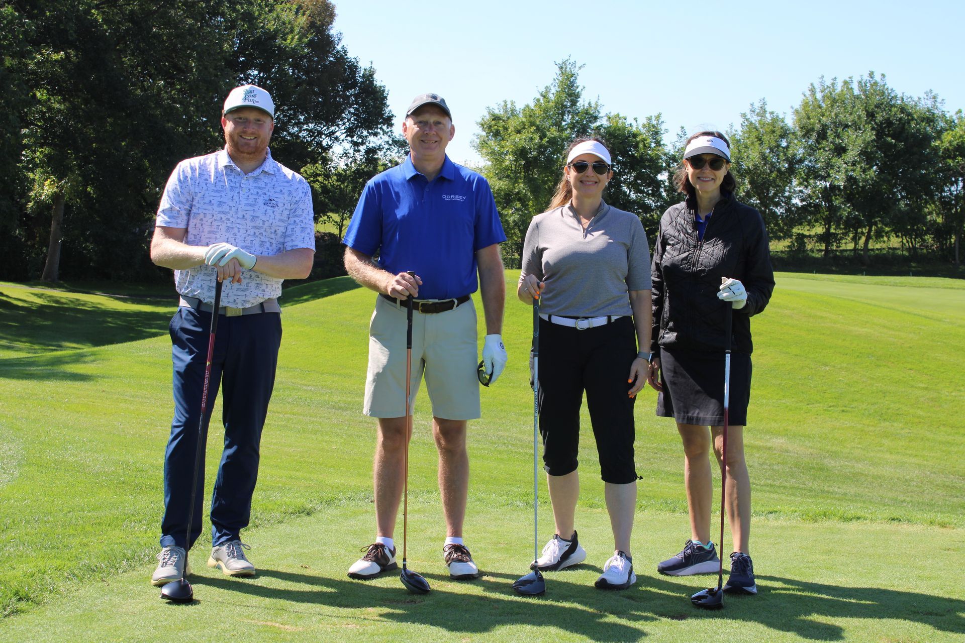 Four people stand on a sunny golf course, each holding a golf club and smiling for a group portrait.