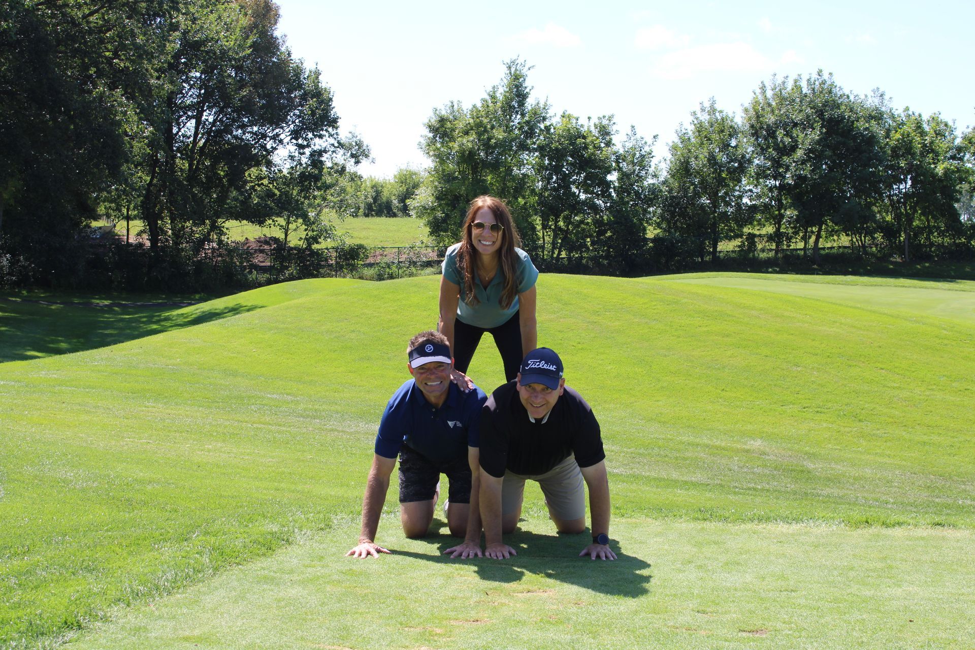 Two people in golf attire on their hands and knees supporting a person standing behind them on a grassy golf course.
