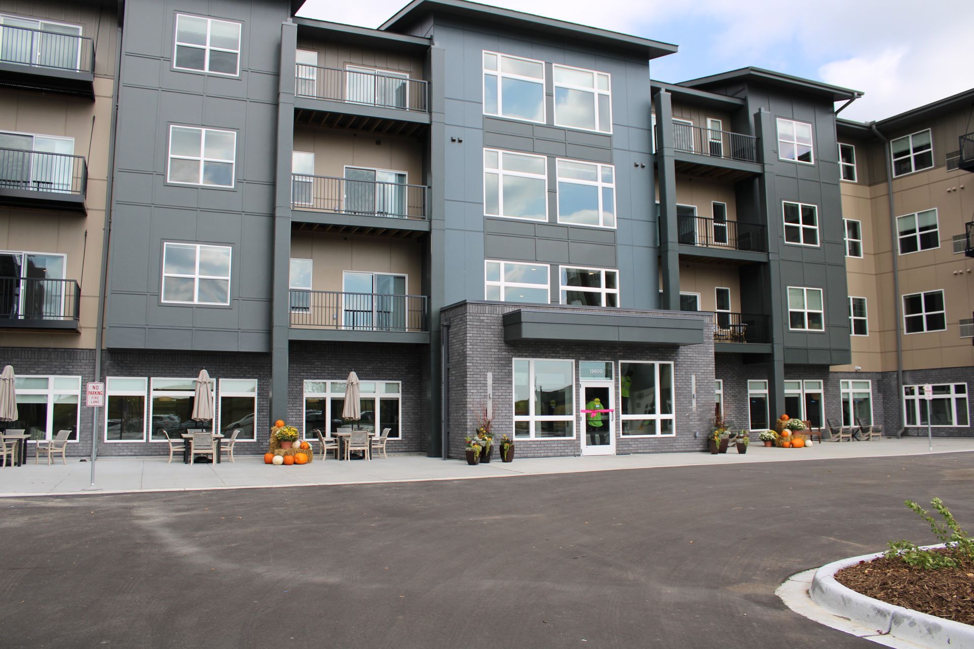 A multi-story modern apartment building featuring gray siding, balconies, large windows, and a ground-level entrance.