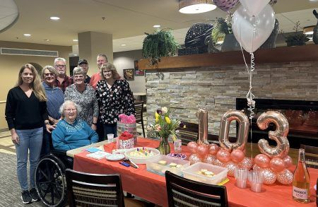A group poses behind a table decorated for a 103rd birthday, featuring rose gold balloons and a cake in a community space.
