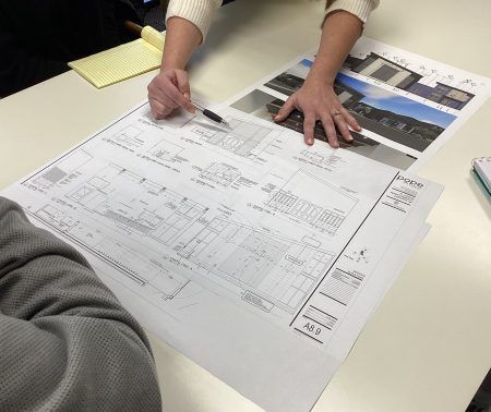 Two pairs of hands review architectural blueprints and building renderings on a white table.