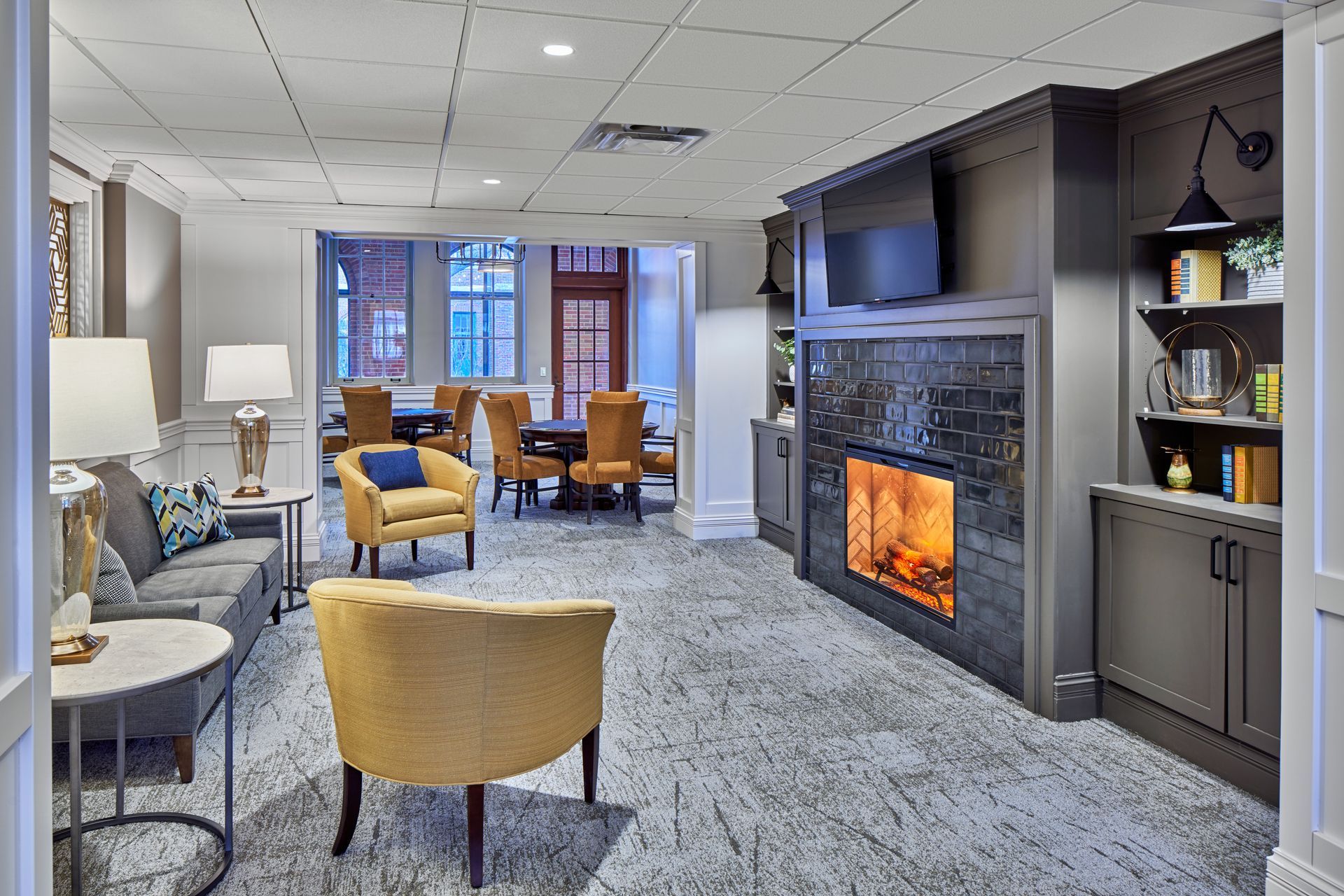 Modern lounge with gray carpet, mustard chairs, wall fireplace, and windows at the far end.