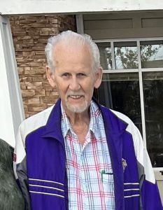 A person with short white hair and a beard, wearing a purple jacket over a plaid shirt, smiling outdoors.